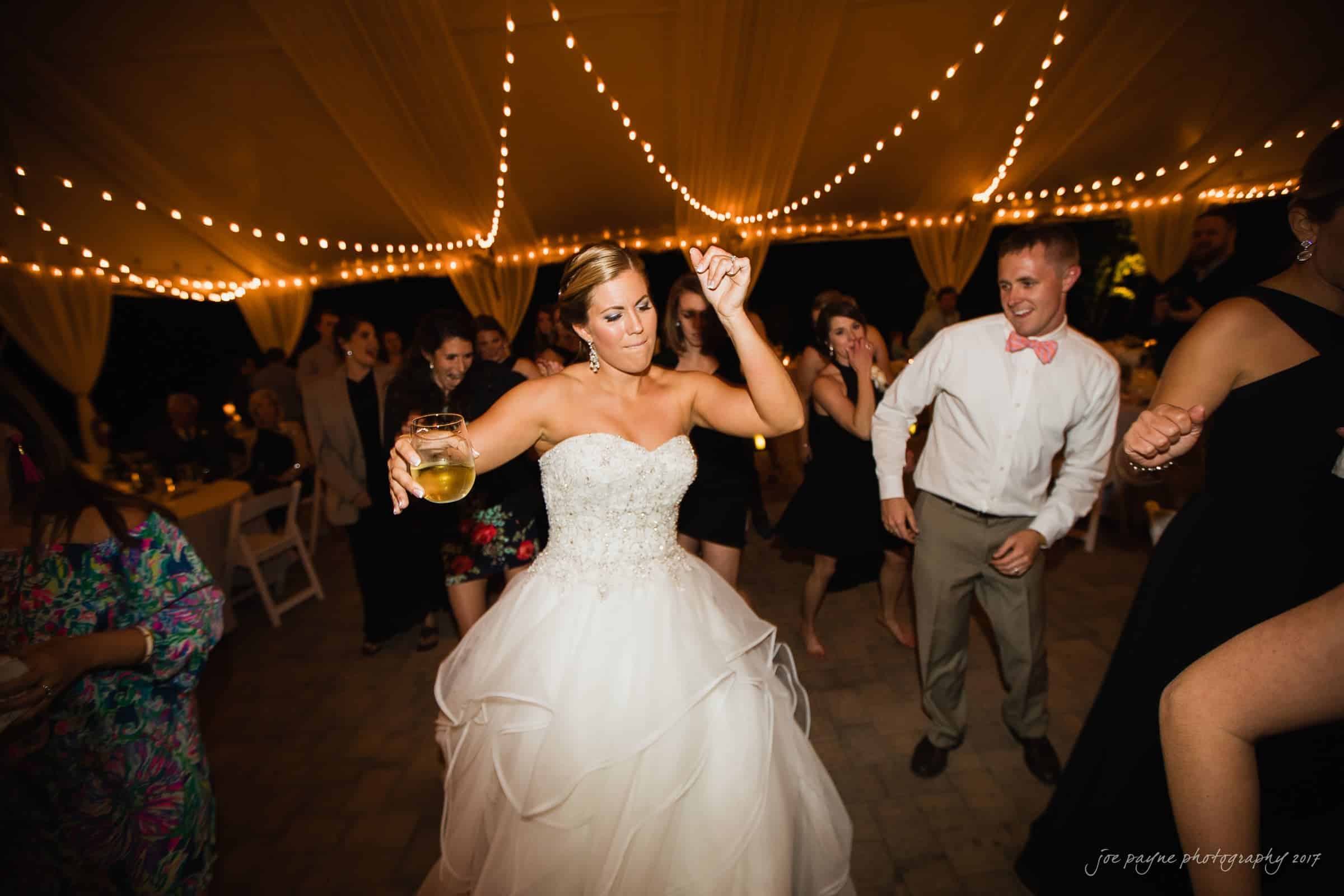 A bride in a white wedding gown dances joyfully under a canopy with string lights. She holds a drink, surrounded by guests in formal attire, all enjoying the celebration.