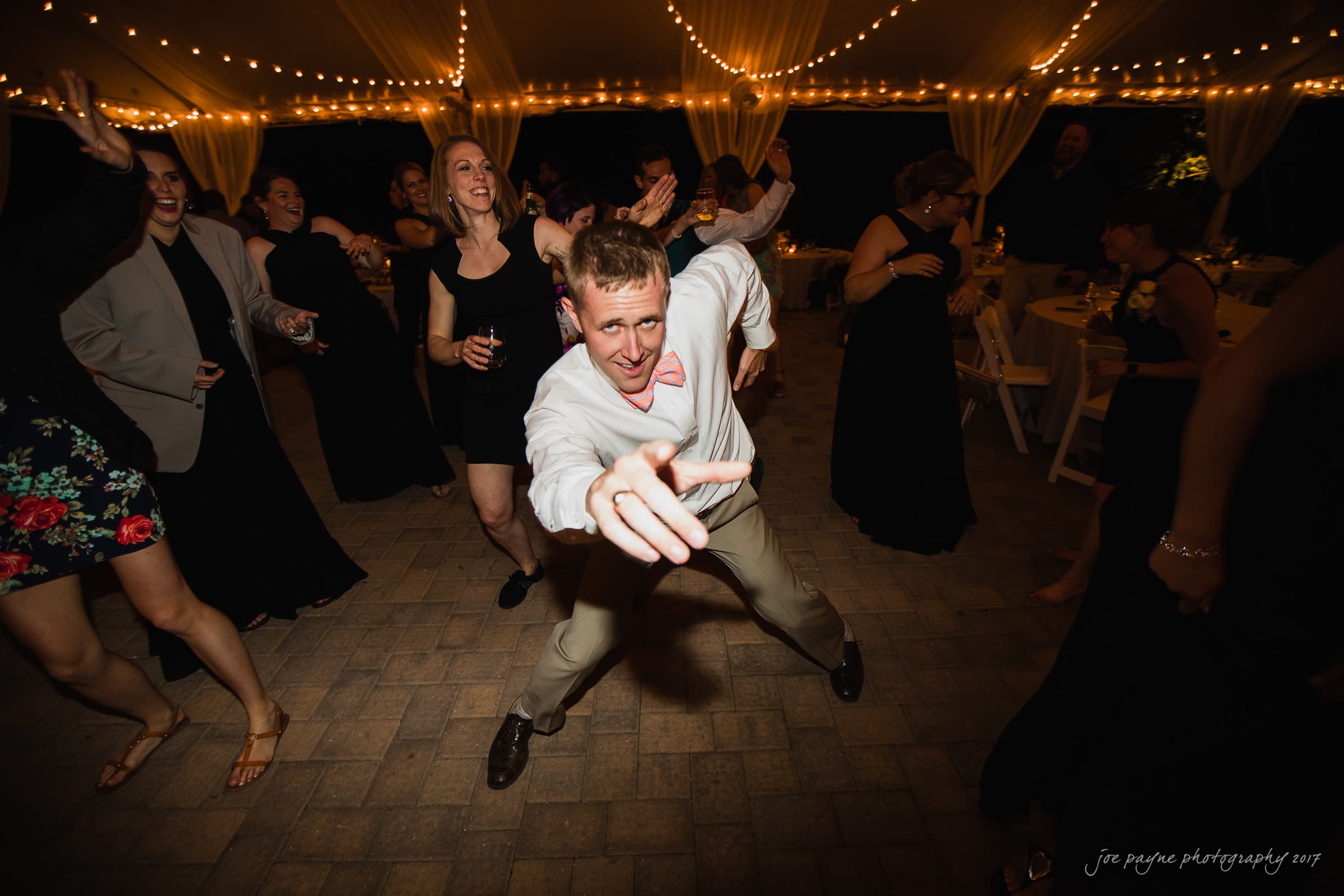 A lively dance floor scene at a party, with a person in a white shirt and pink bow tie enthusiastically pointing towards the camera. String lights and people dancing fill the background, creating a joyful and energetic atmosphere.