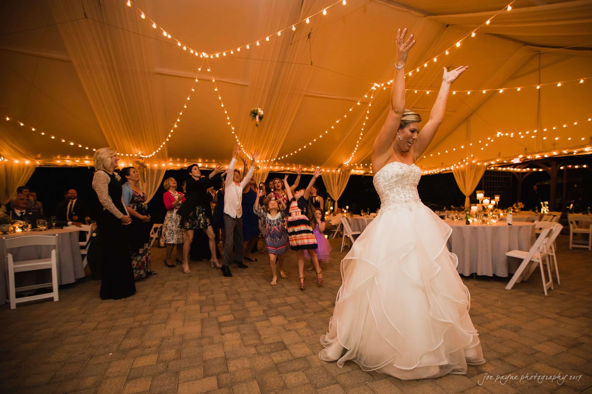 A bride in a white gown joyfully throws her bouquet over her shoulder at a night wedding reception. Guests eagerly reach to catch it under a tent decorated with string lights, surrounded by tables and chairs.