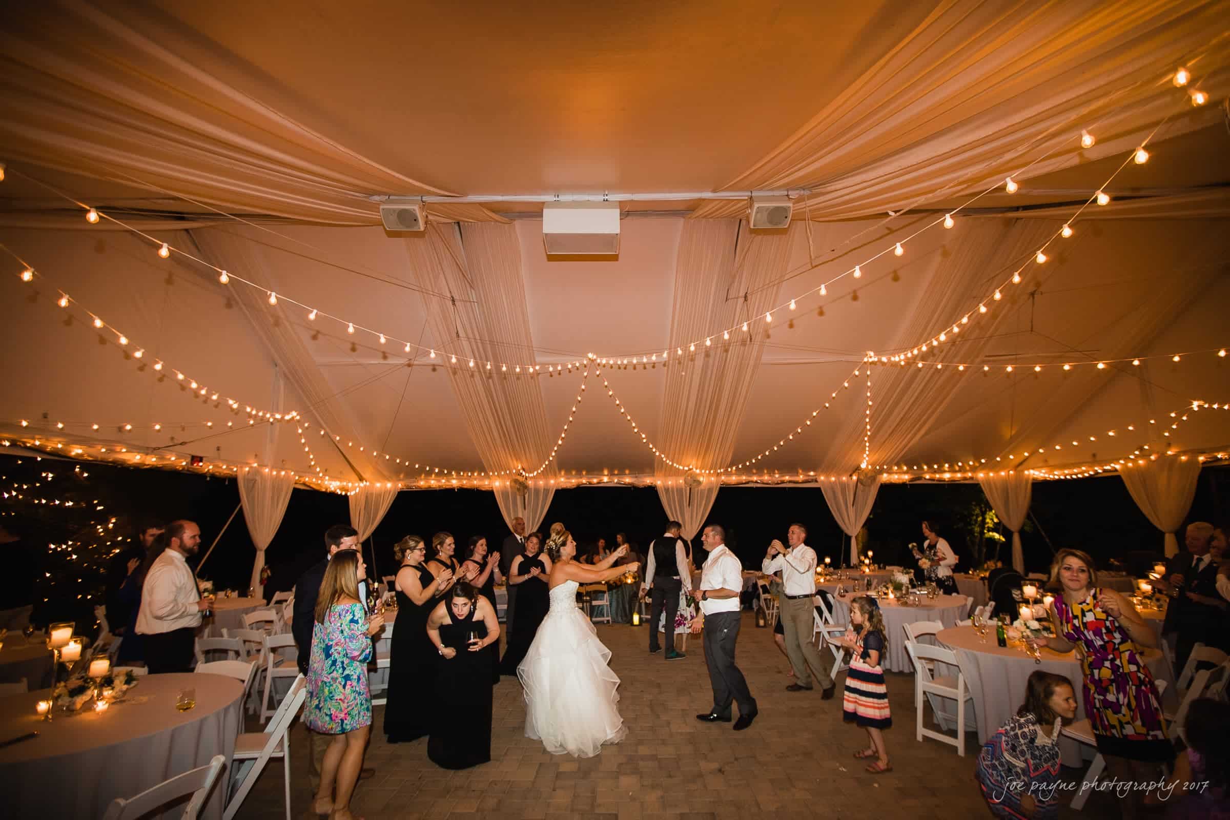 A bride dances with a man under a beautifully lit canopy at an outdoor wedding reception. Guests are mingling and dancing around them, with tables decorated with candles and string lights creating a warm, festive atmosphere.