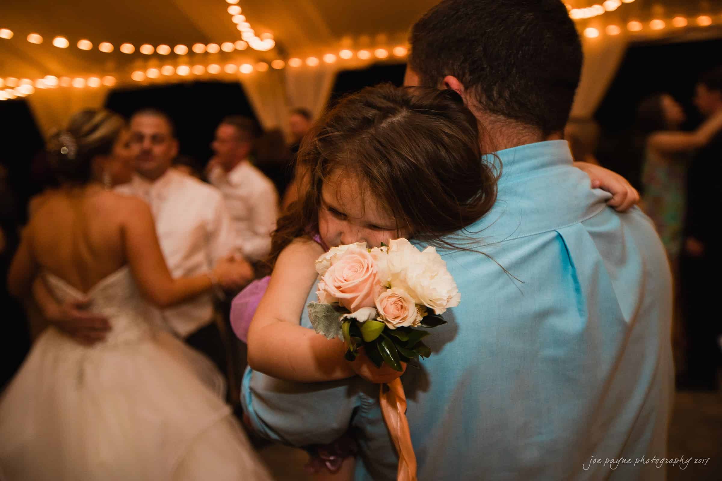 A man in a blue shirt holds a young girl wearing a purple dress who is holding a bouquet of flowers. They are at an indoor event with string lights above. In the background, there are other people, including a bride in a wedding gown.