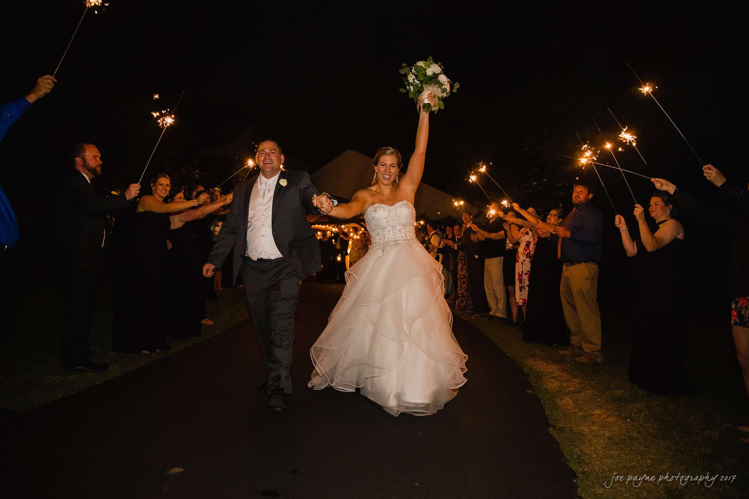 A joyful bride in a white gown and groom in a suit walk hand in hand down a path at night, surrounded by guests holding sparklers. The bride holds a bouquet aloft, and both are smiling brightly.