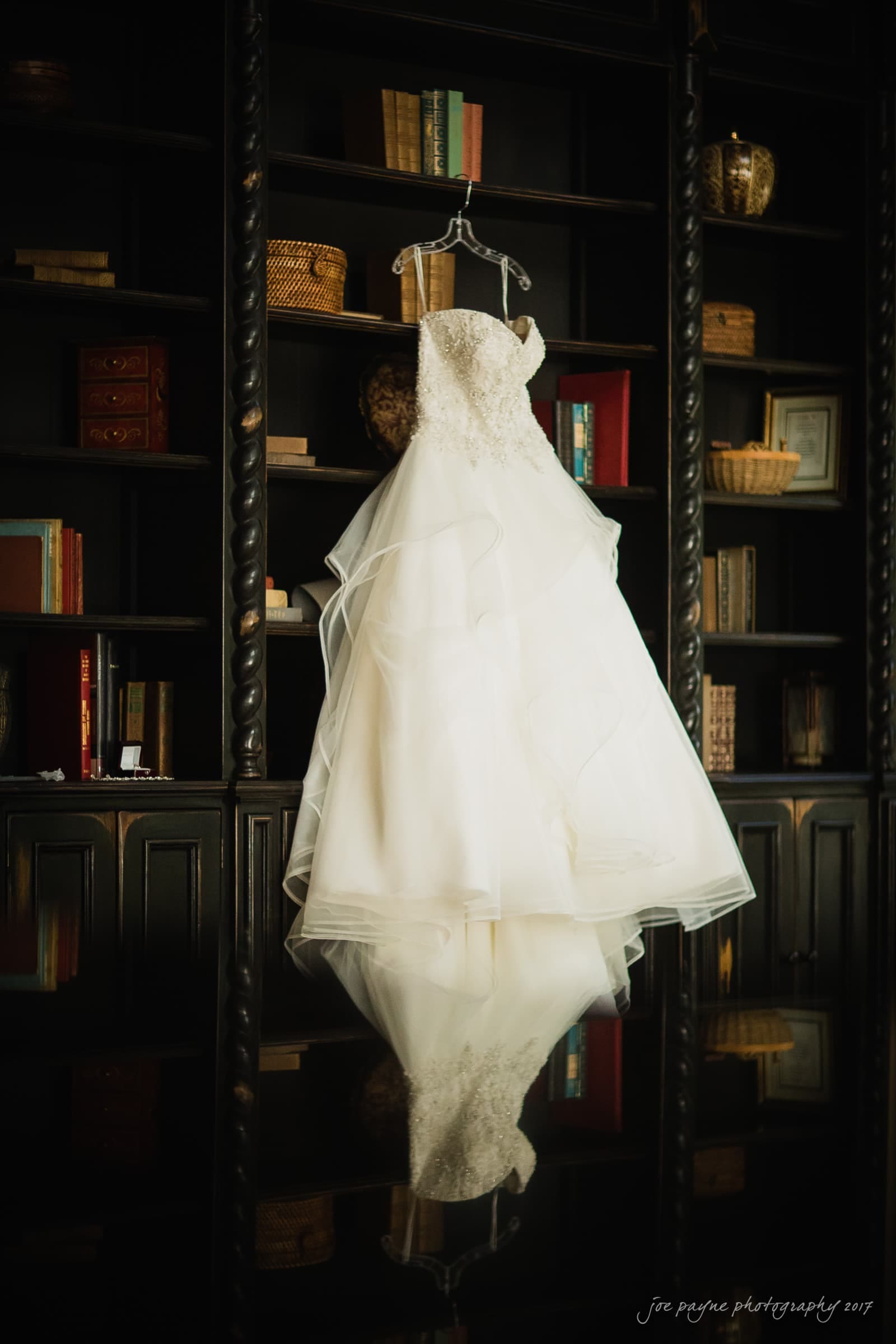 A white wedding dress with intricate lace detail hangs elegantly against a dark wooden bookshelf filled with various books and baskets, creating a striking contrast.