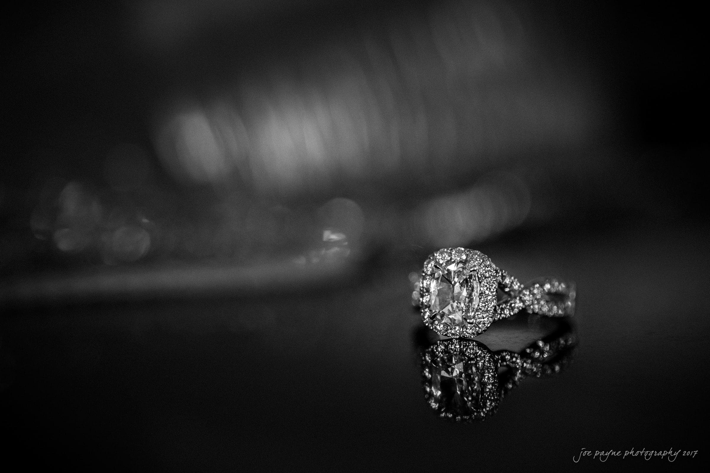 A black and white photo of an ornate diamond ring with intricate detailing, resting on a reflective surface, creating a soft reflection. The background is blurred, emphasizing the rings elegance and shine.