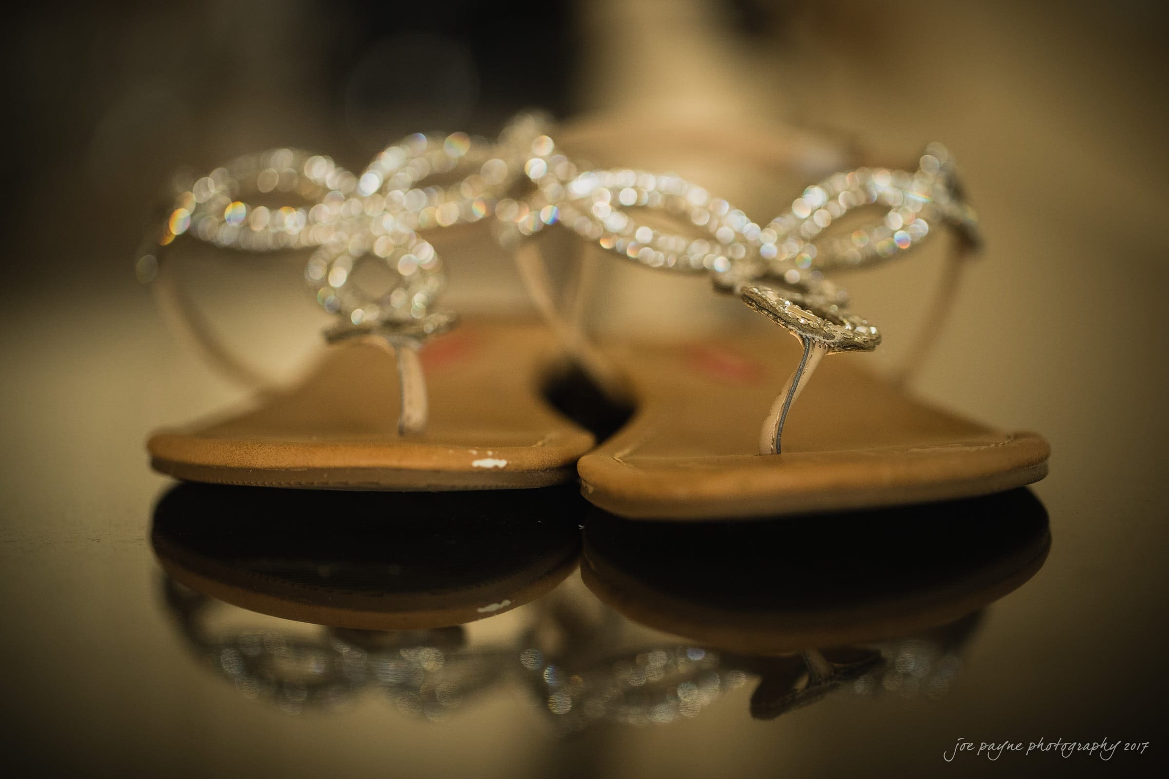 Close-up of elegant flat sandals with intricate silver embellishments on a reflective surface. The focus is on the shimmering details of the straps.