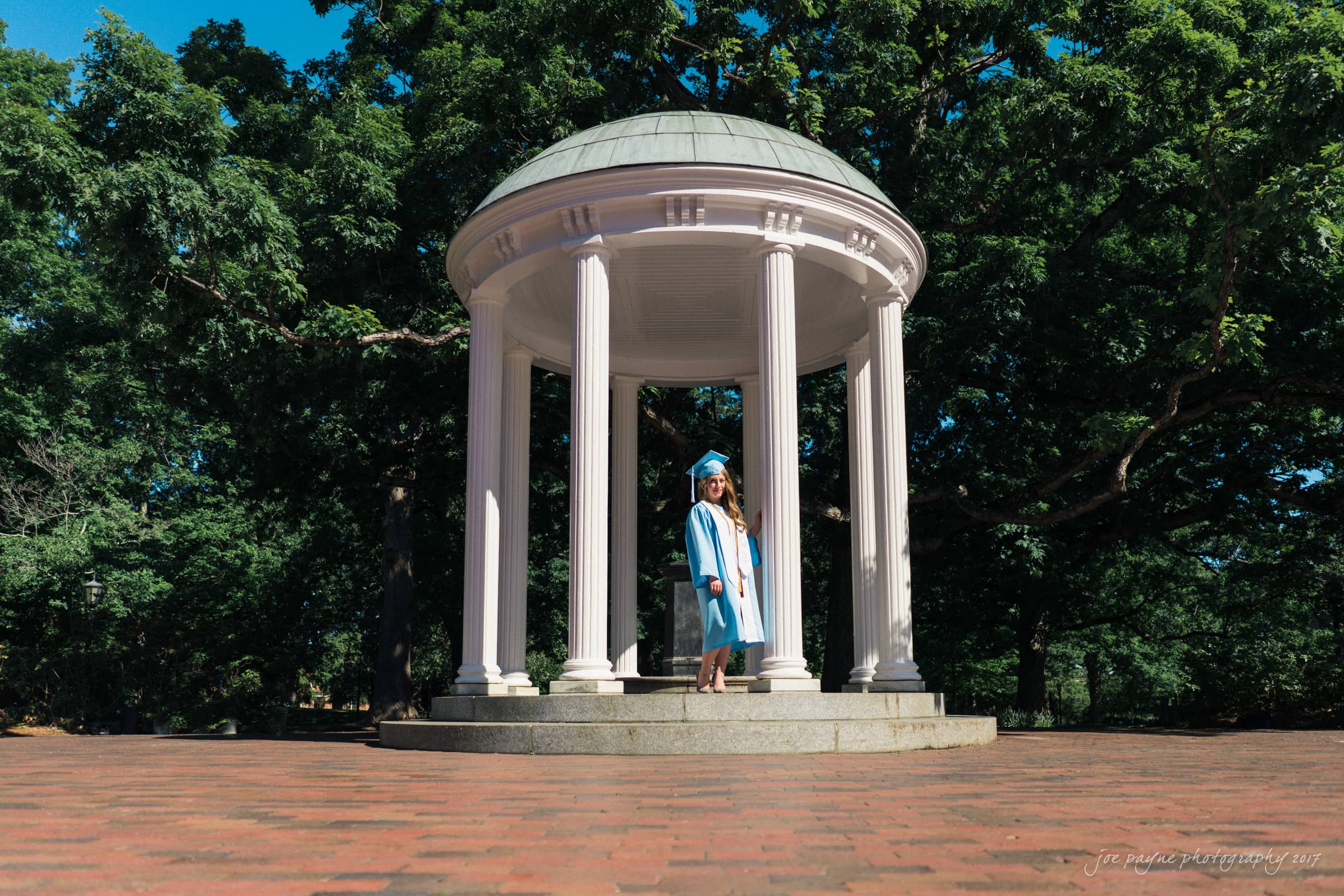 A graduate in a blue cap and gown poses under a small, white domed pavilion with tall columns, surrounded by lush green trees and set on a brick platform. The sky is clear and blue.