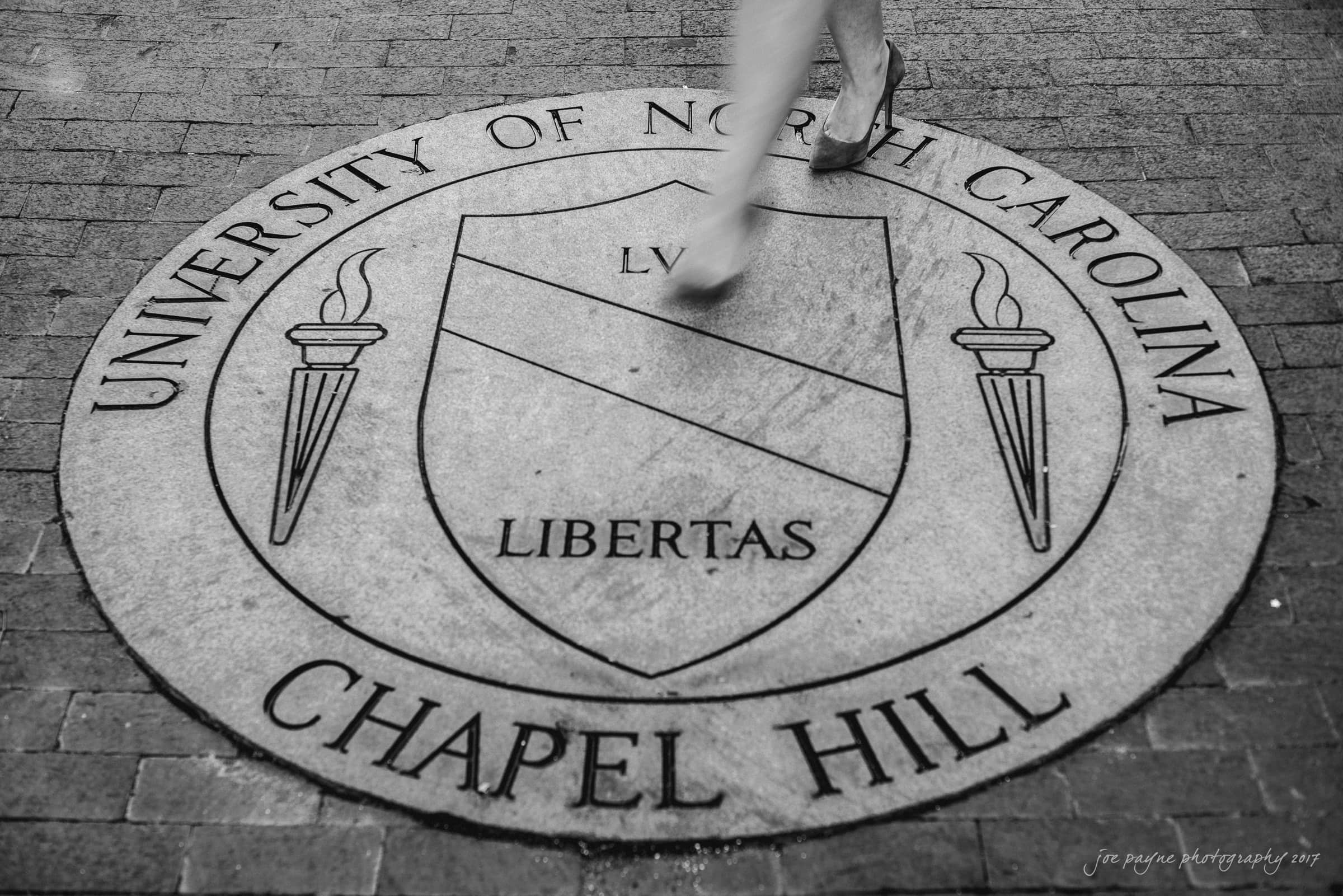 A person walks over a circular stone emblem on a brick path, displaying the University of North Carolina Chapel Hill seal with Libertas and two torches. The image is in black and white.