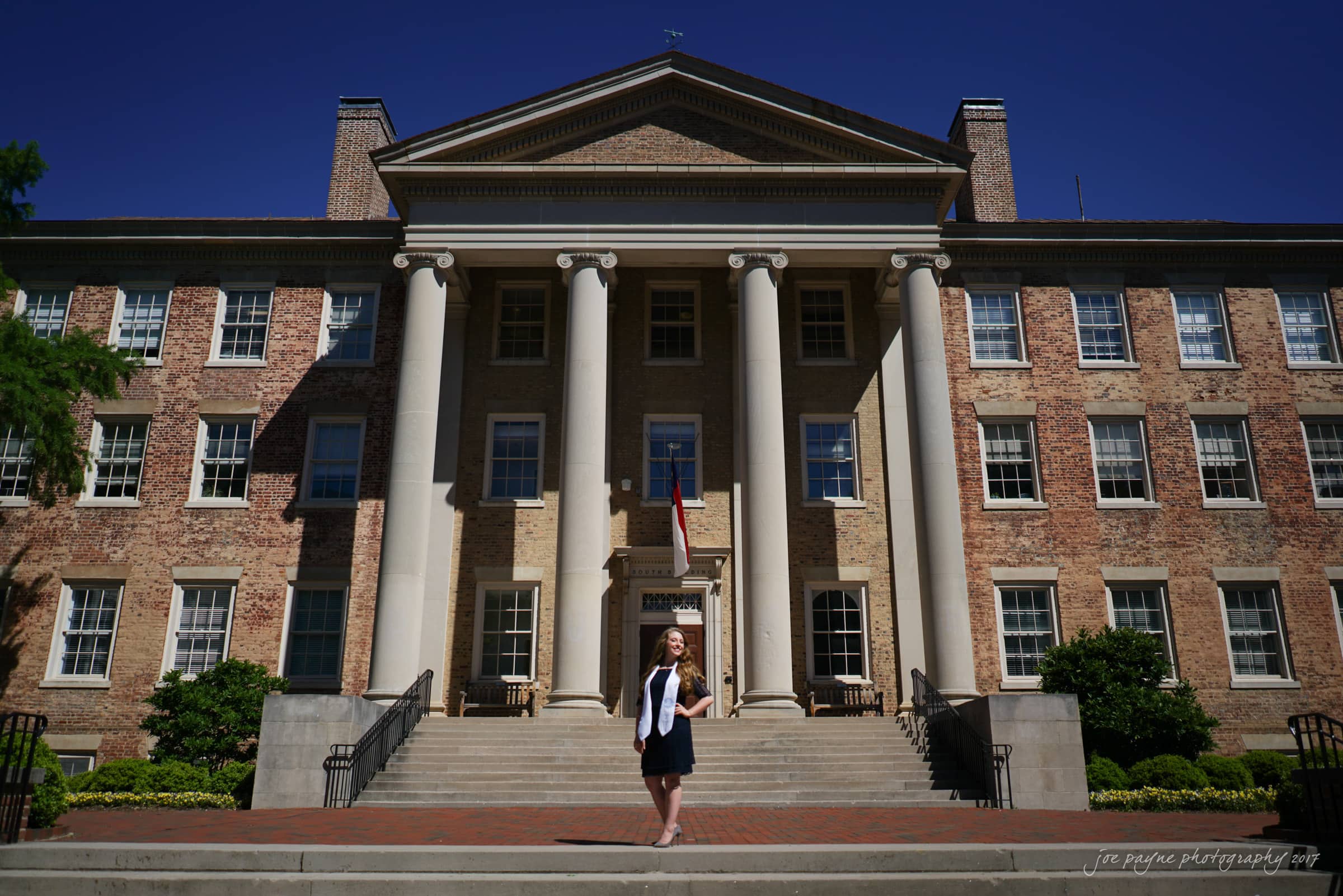 A person stands on the steps of a large brick building with tall columns and a flag in the center. The building has multiple windows and is set against a clear blue sky. Greenery flanks the structure.