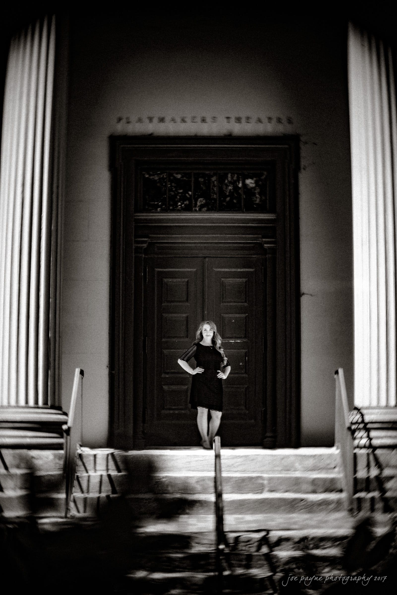 Black and white photo of a person in a dark dress standing confidently in front of the large doors of Playmakers Theater. They are flanked by tall, white columns. The overall mood is dramatic and timeless.