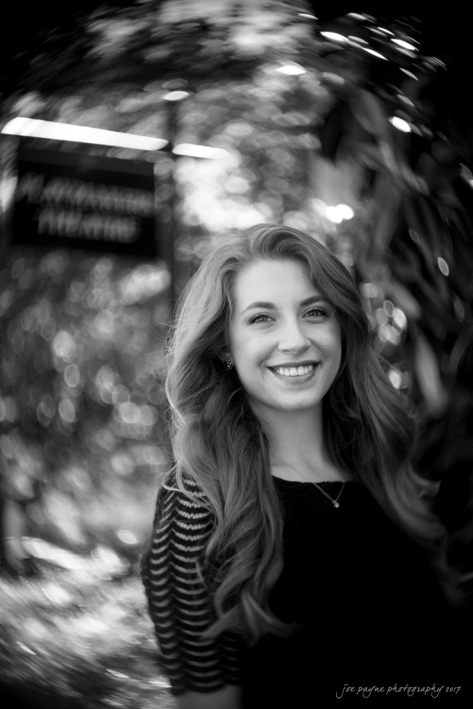 A woman with long hair smiles at the camera in a black and white photo. The background features swirling bokeh and blurred leaves, creating a dreamy effect. A sign with illegible text is partially visible in the background.