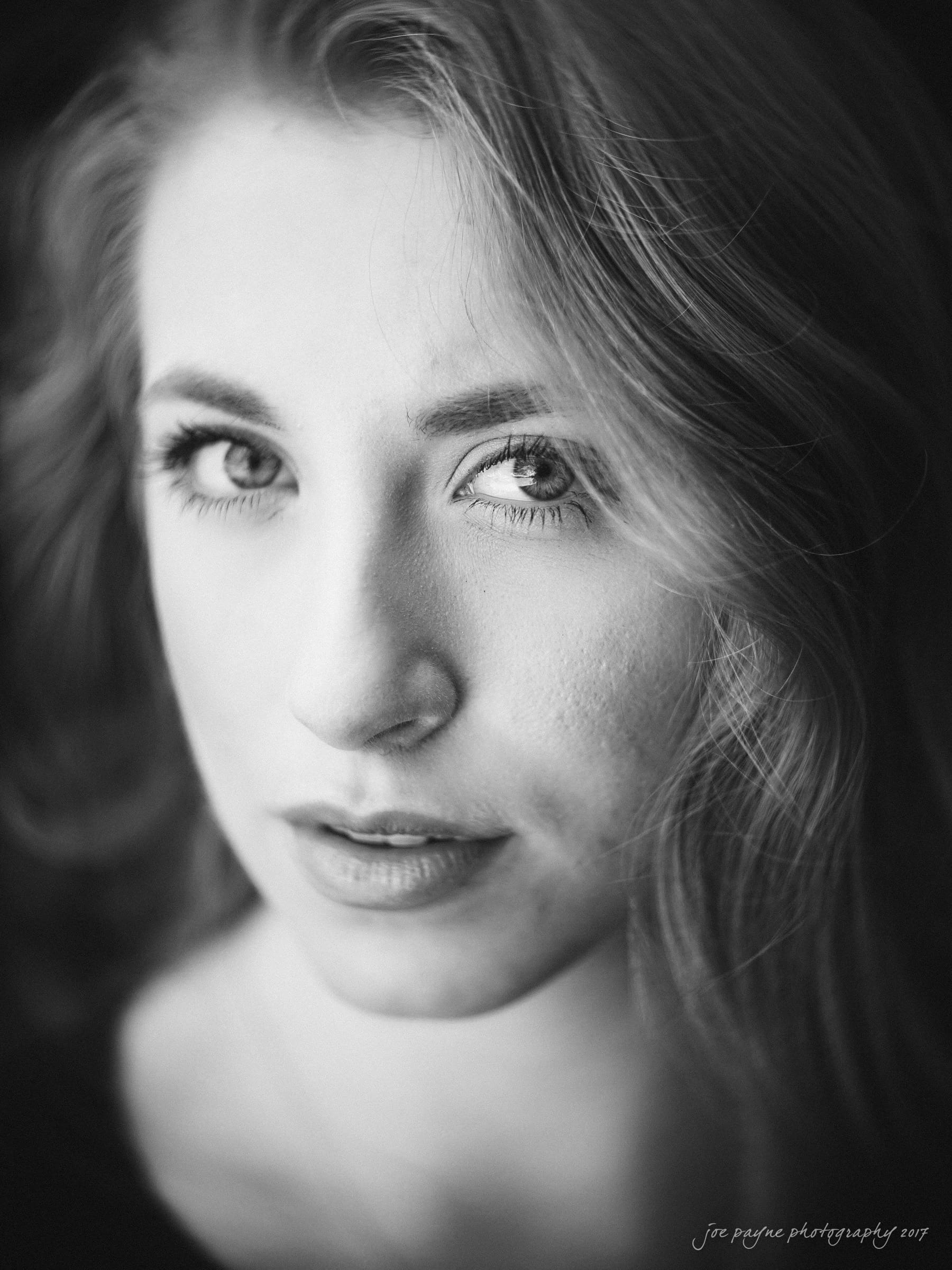 Black and white close-up portrait of a person with long hair, looking directly at the camera with a serious expression. The image is taken from a slight angle, highlighting facial features and texture.