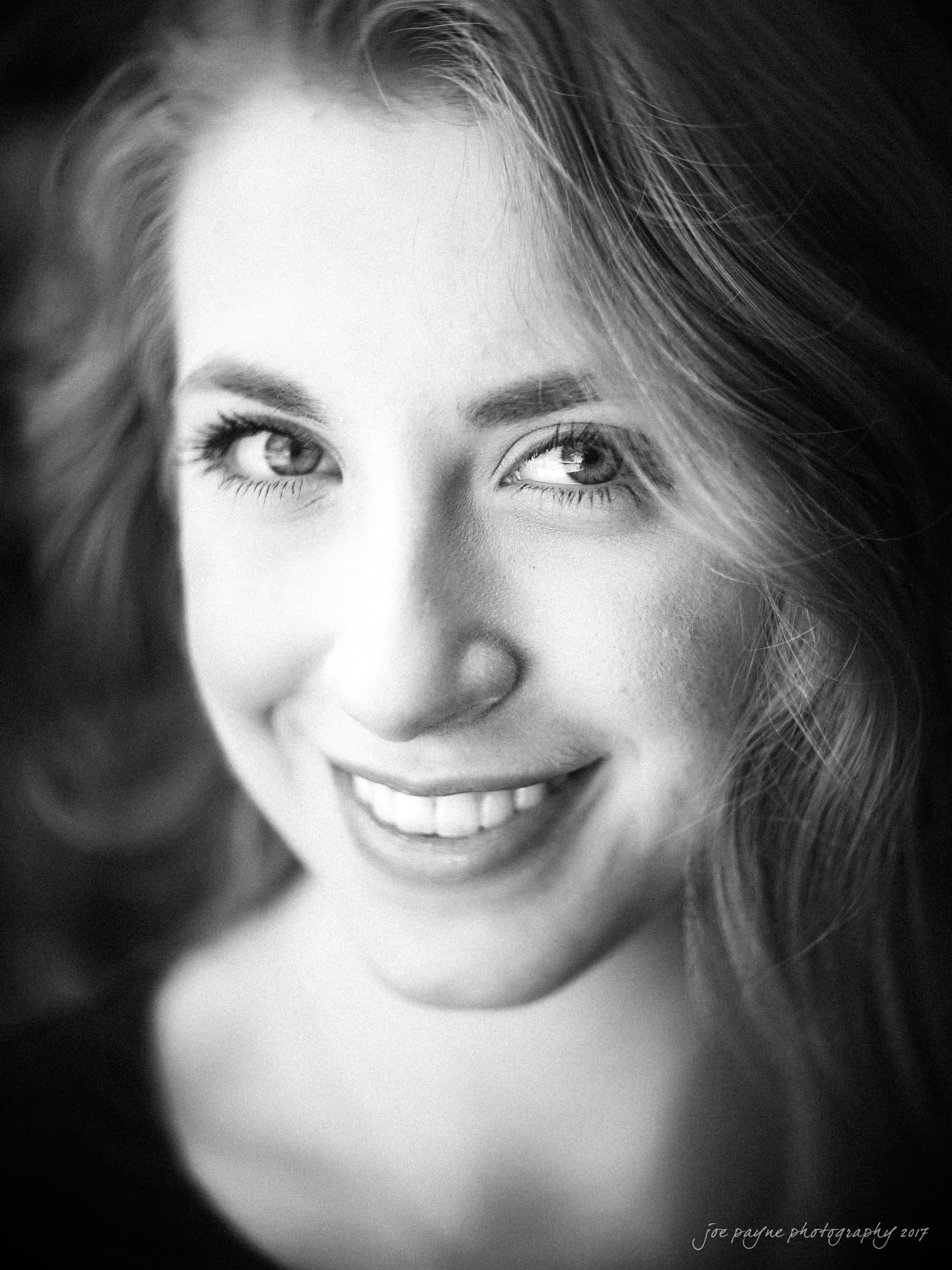 Black and white portrait of a person with long hair, smiling warmly. The lighting highlights their eyes and creates soft shadows on their face. The background is blurred, drawing focus to the subjects expression.