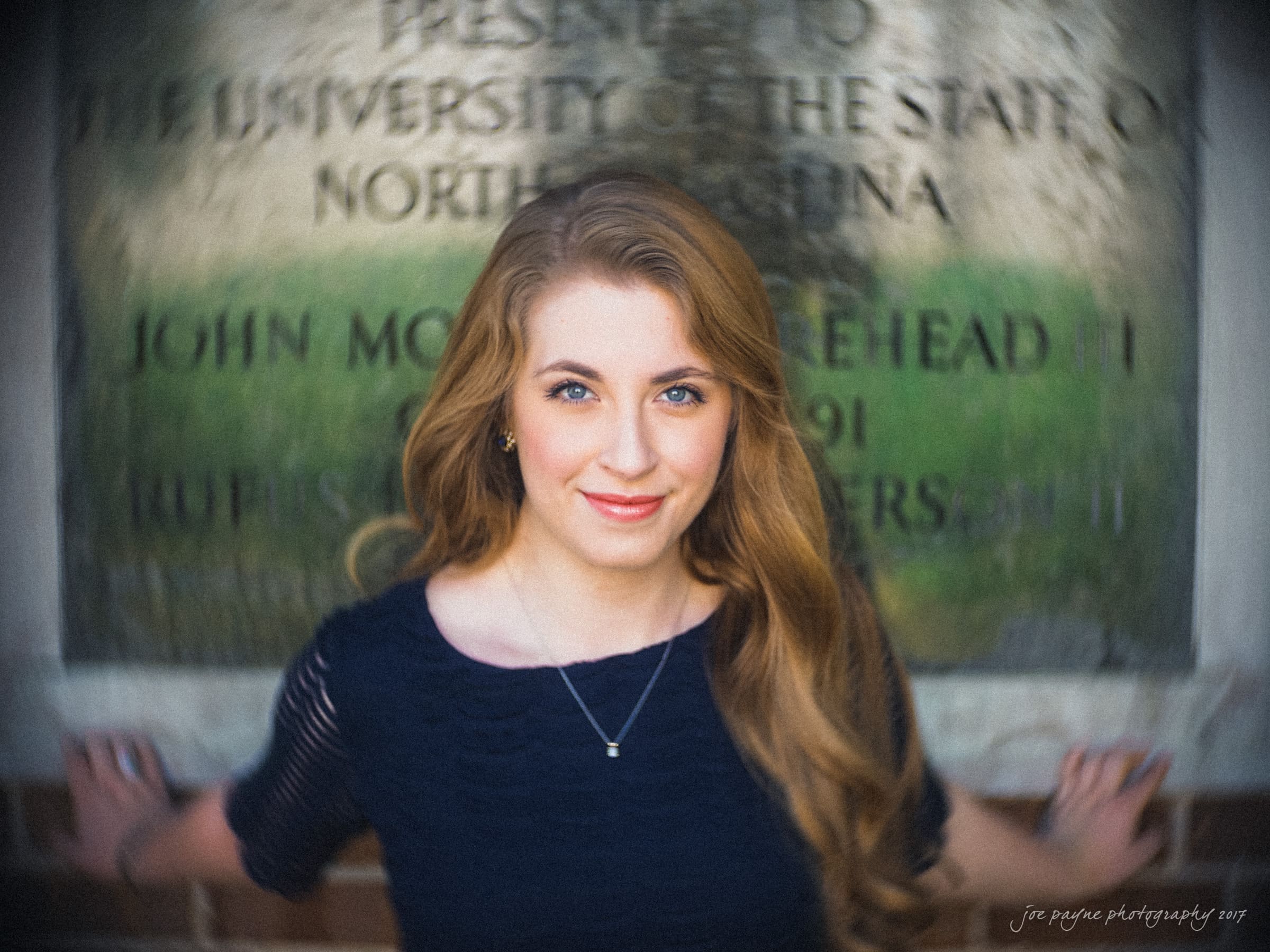 A person with long, wavy hair and a necklace stands in front of a blurred background featuring a plaque with text. They are wearing a dark blue top and are smiling softly at the camera.