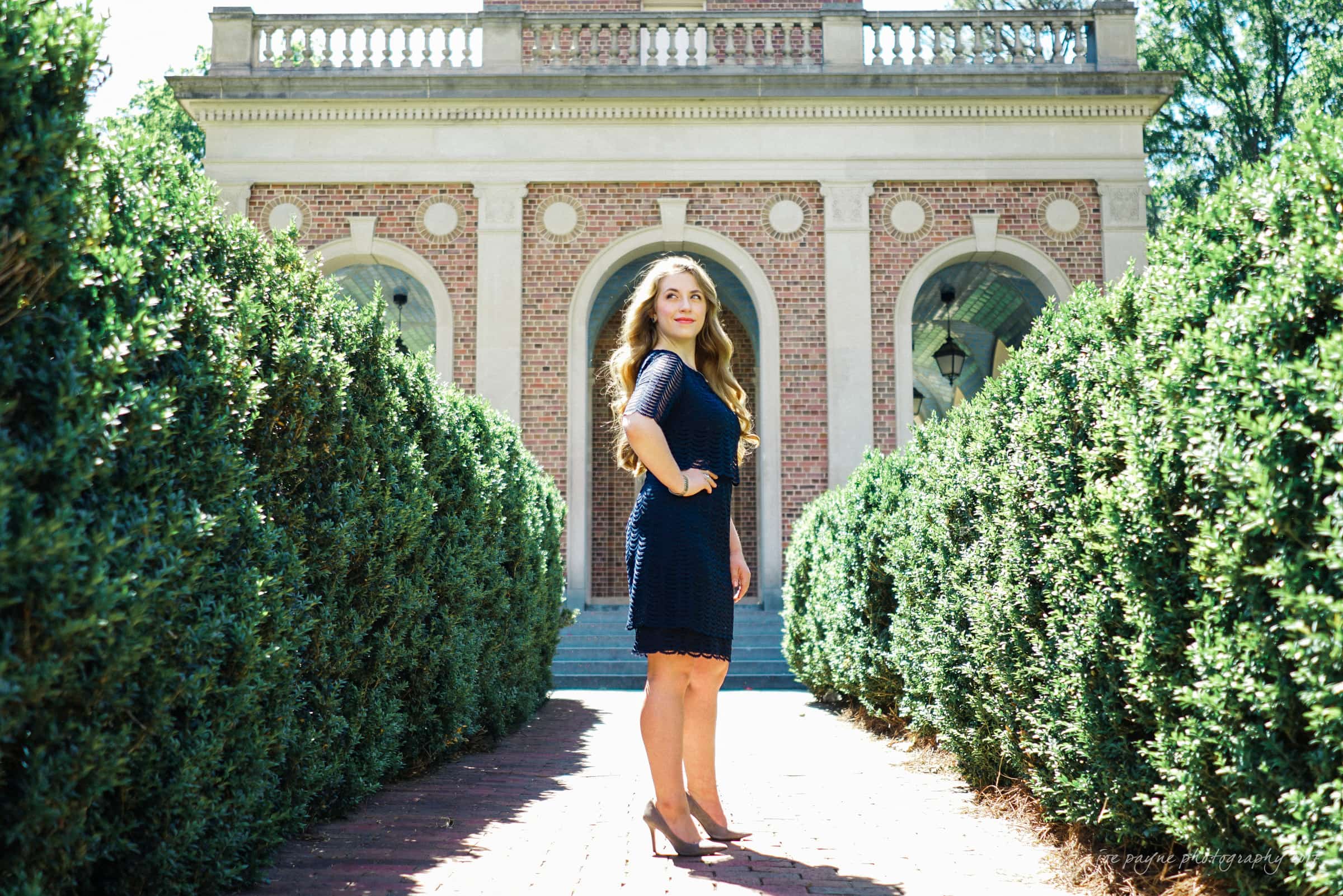 A woman in a blue dress stands confidently on a brick pathway lined with lush green hedges. Behind her is a brick building with three arched doorways and large windows, capturing a sunny day.