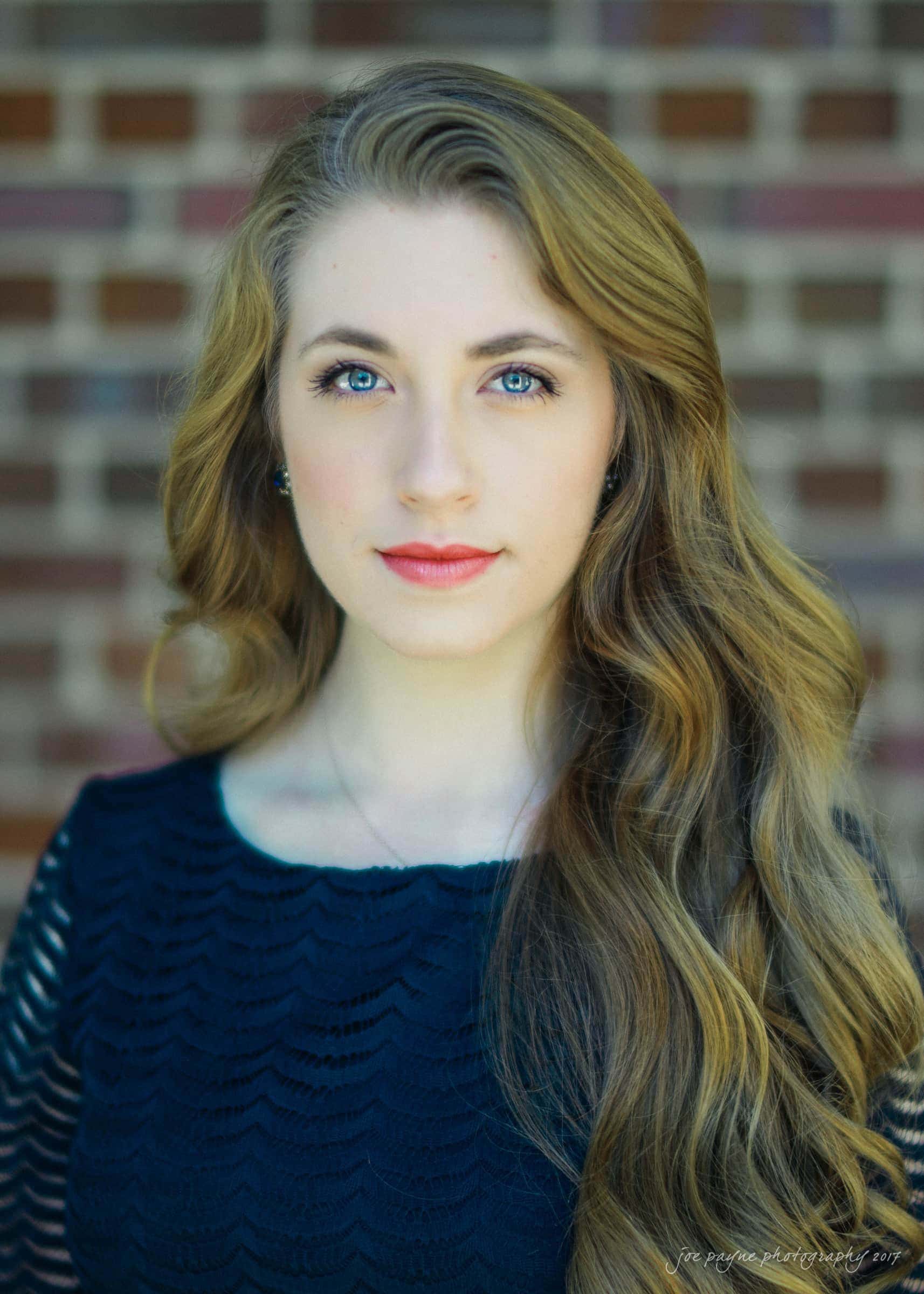 A woman with long, wavy hair and a serious expression stands in front of a brick wall. She is wearing a dark, textured top and has bright blue eyes. Her hair is cascading over one shoulder.