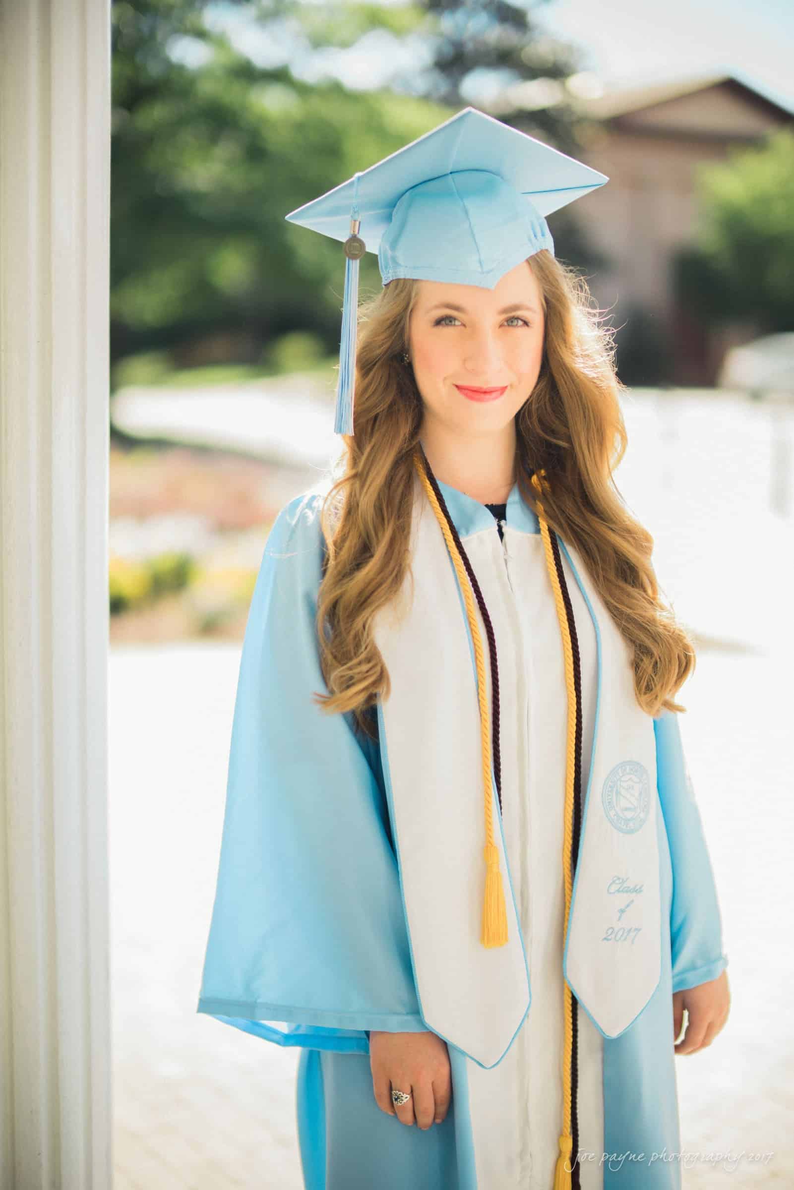 A graduate in a light blue cap and gown stands outdoors. She is wearing a white stole and honor cords. Sunlight shines on her as she smiles at the camera.