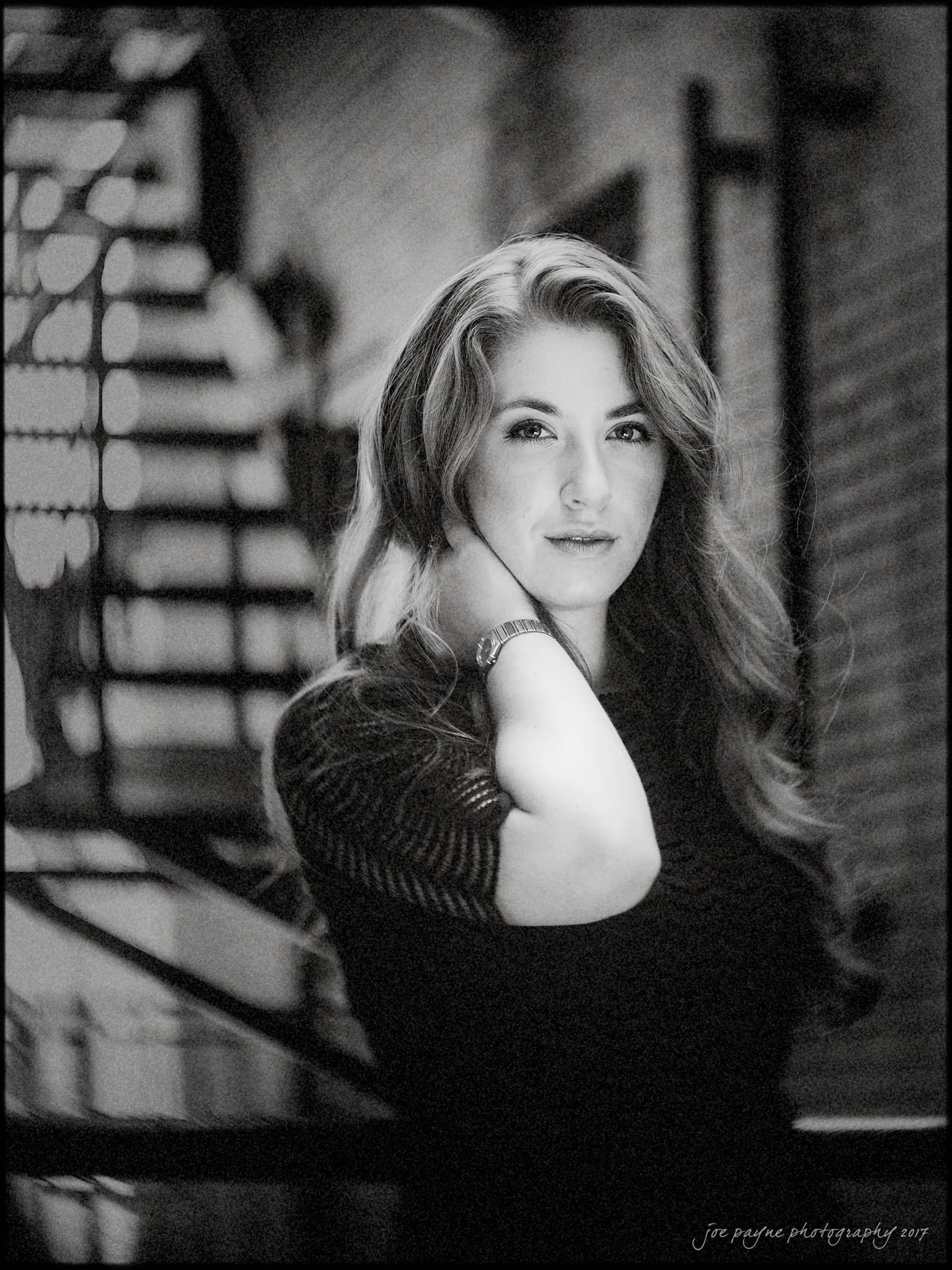 A black and white portrait of a woman with long hair in a dark, striped top. She stands in front of a blurred background with stairs, gently holding her neck with one hand and looking towards the camera, conveying a calm expression.