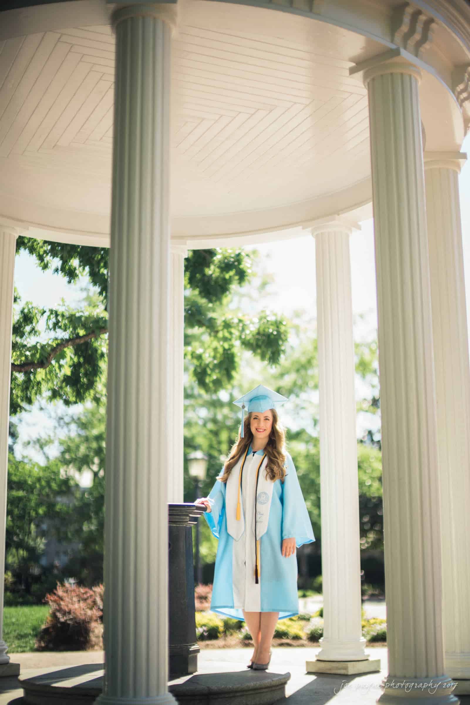 A graduate in a light blue cap and gown stands smiling beneath a white gazebo with columns, surrounded by greenery. She holds several cords and stoles, indicating academic achievements.