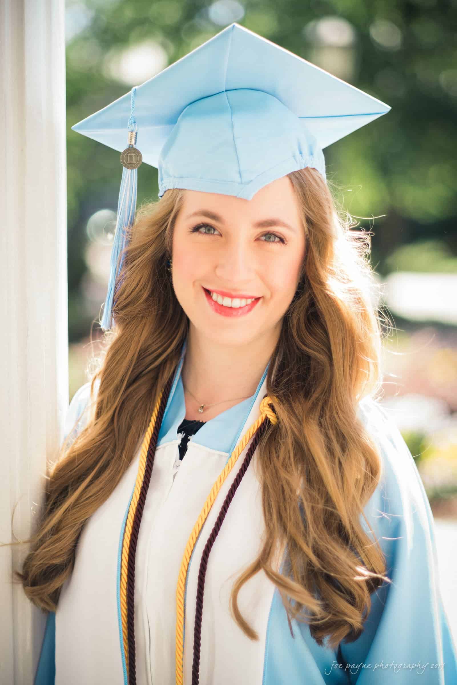 A graduate wearing a light blue cap and gown smiles brightly. They have long, wavy hair and are standing outdoors, with a blurred green background. Multicolored cords hang around their neck.