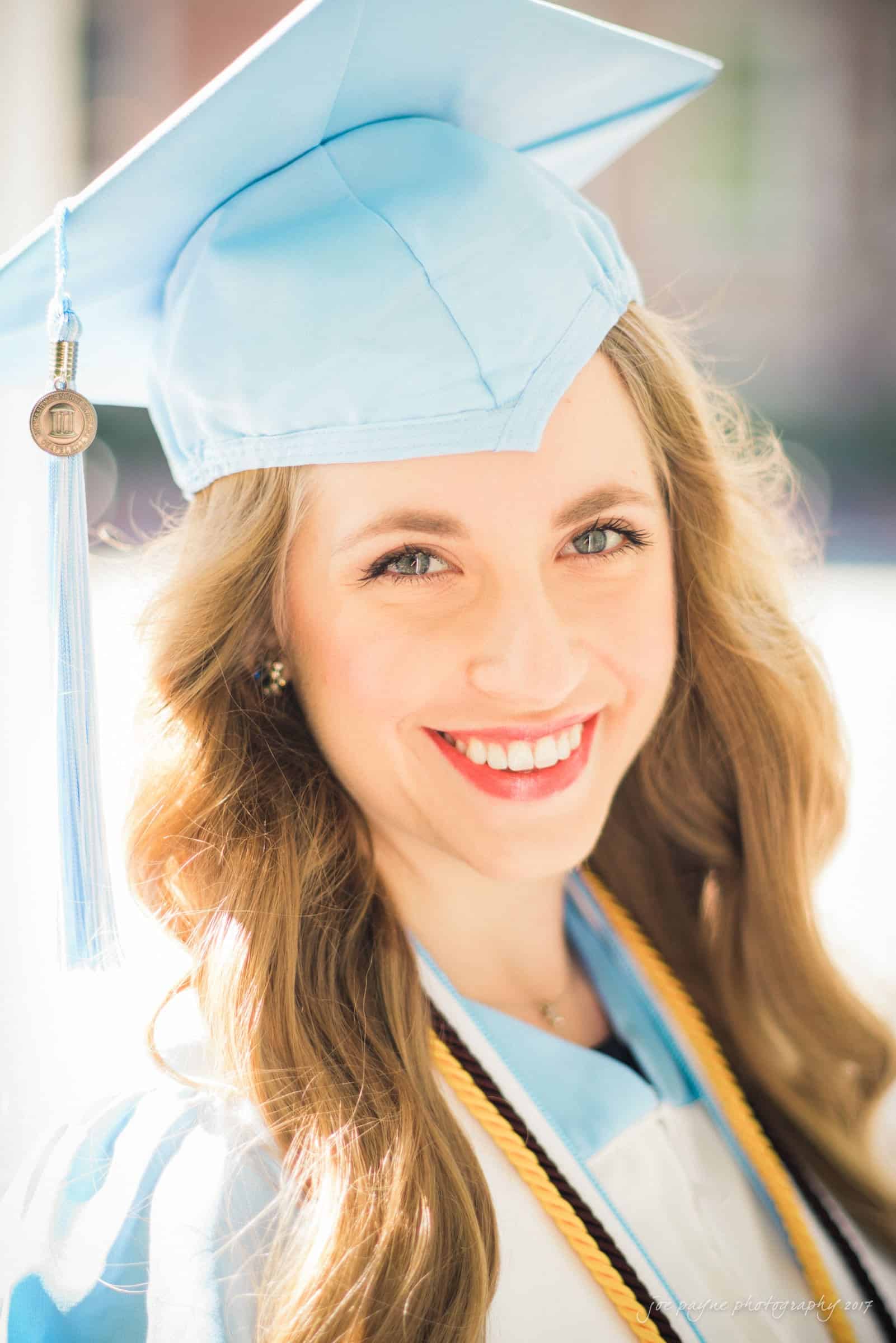 A person in a light blue graduation cap and gown smiles brightly. They have long, wavy hair and wear a gold honor cord. The background is softly blurred, suggesting an outdoor setting.