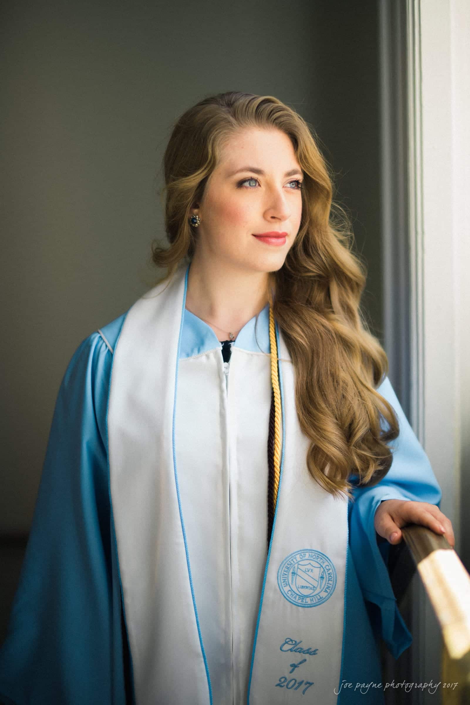 A woman wearing a blue graduation gown and a white stole is looking out of a window. The stole reads Class of 2017 and has an emblem. She has long, wavy hair and a thoughtful expression.