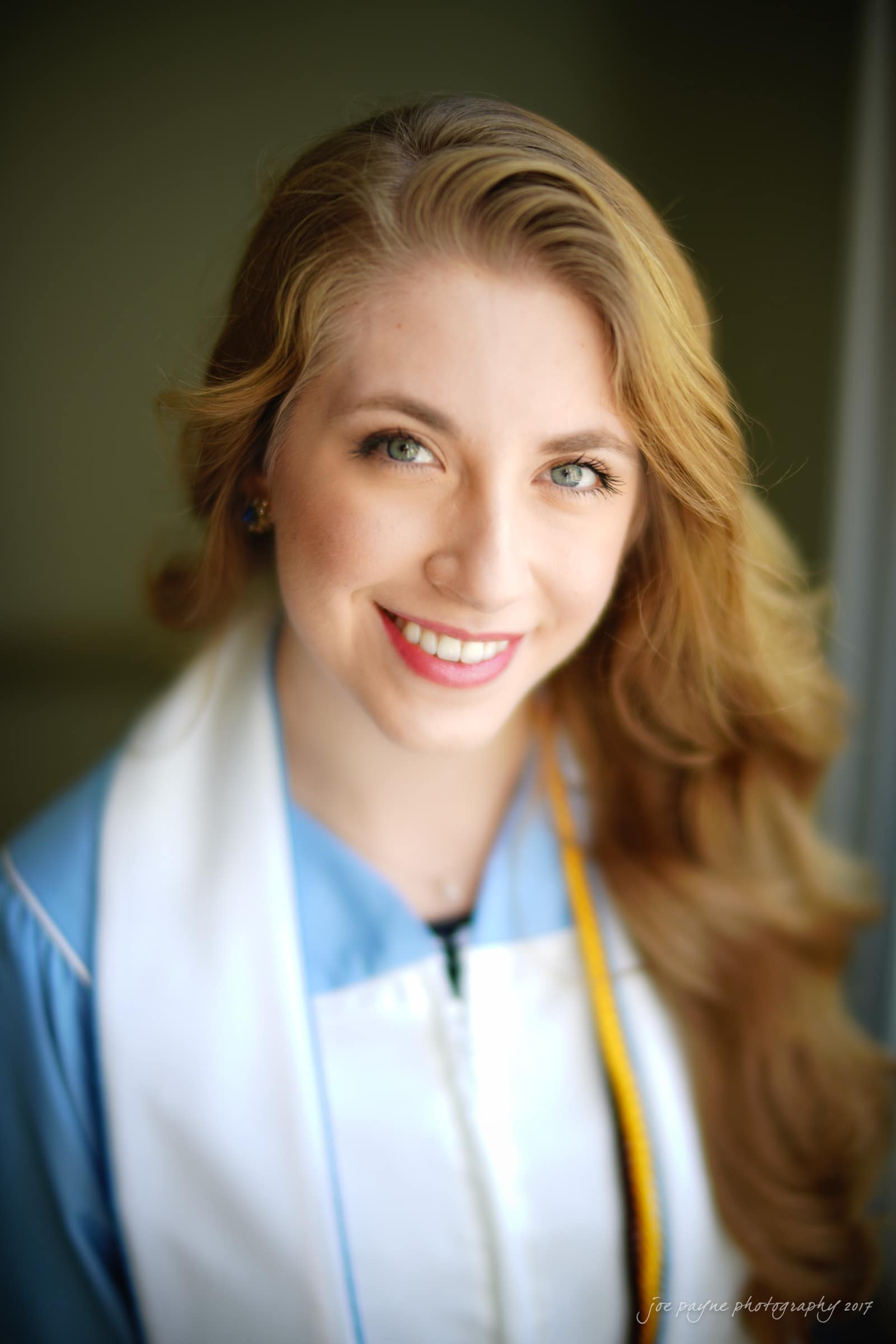 A smiling woman with long, wavy hair wears a light blue graduation gown and white stole, standing against a blurred background.
