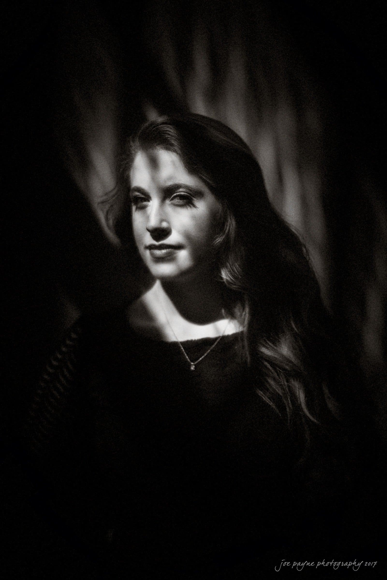 A black and white portrait of a woman with long hair, illuminated by dappled light and shadows. Shes looking slightly to the side with a serene expression. The background is blurred, emphasizing her face and the play of light.