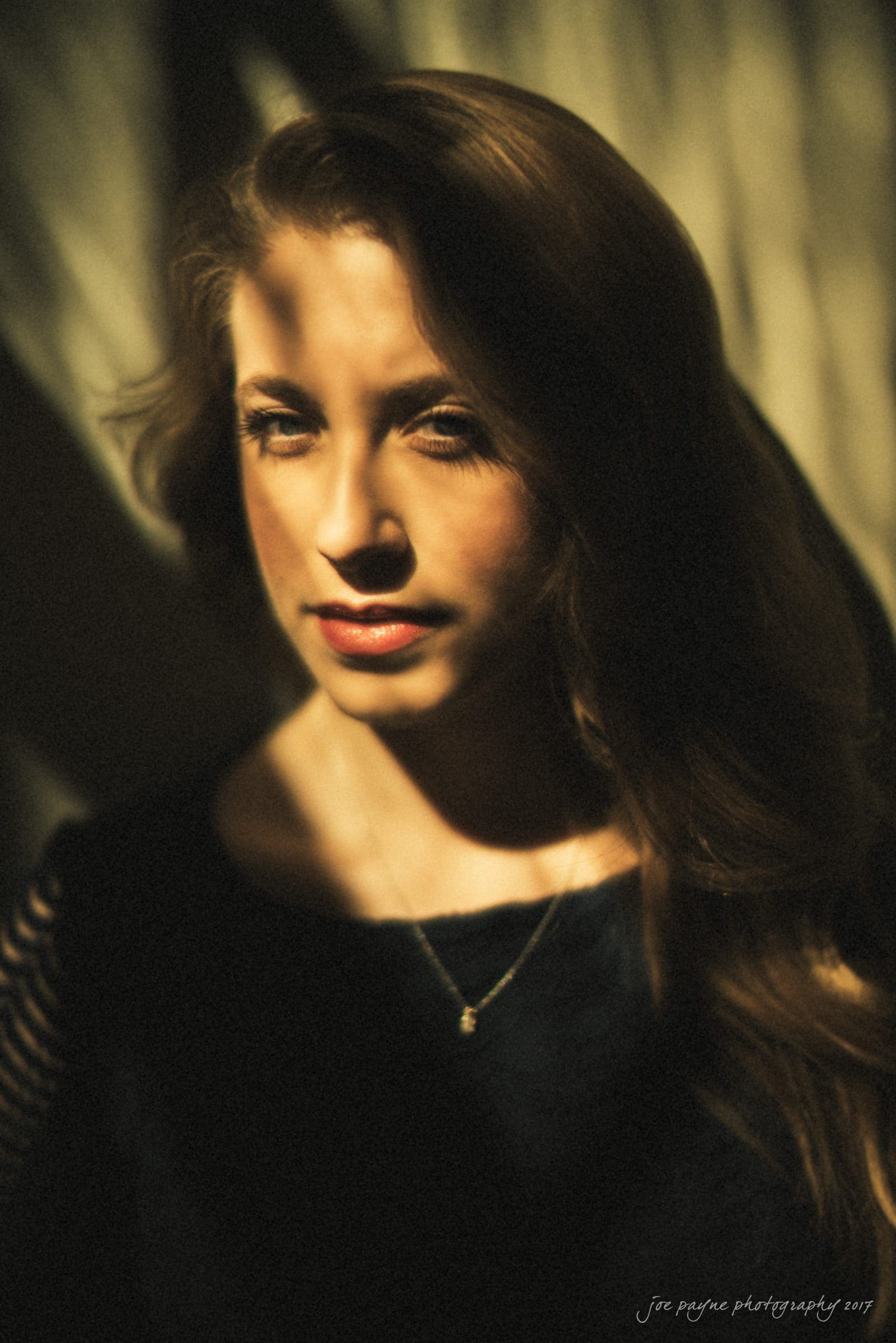 Portrait of a woman with long hair, softly lit with dappled shadows across her face. She is wearing a dark top and a necklace, looking directly at the camera. The background is blurred, creating a warm and introspective atmosphere.