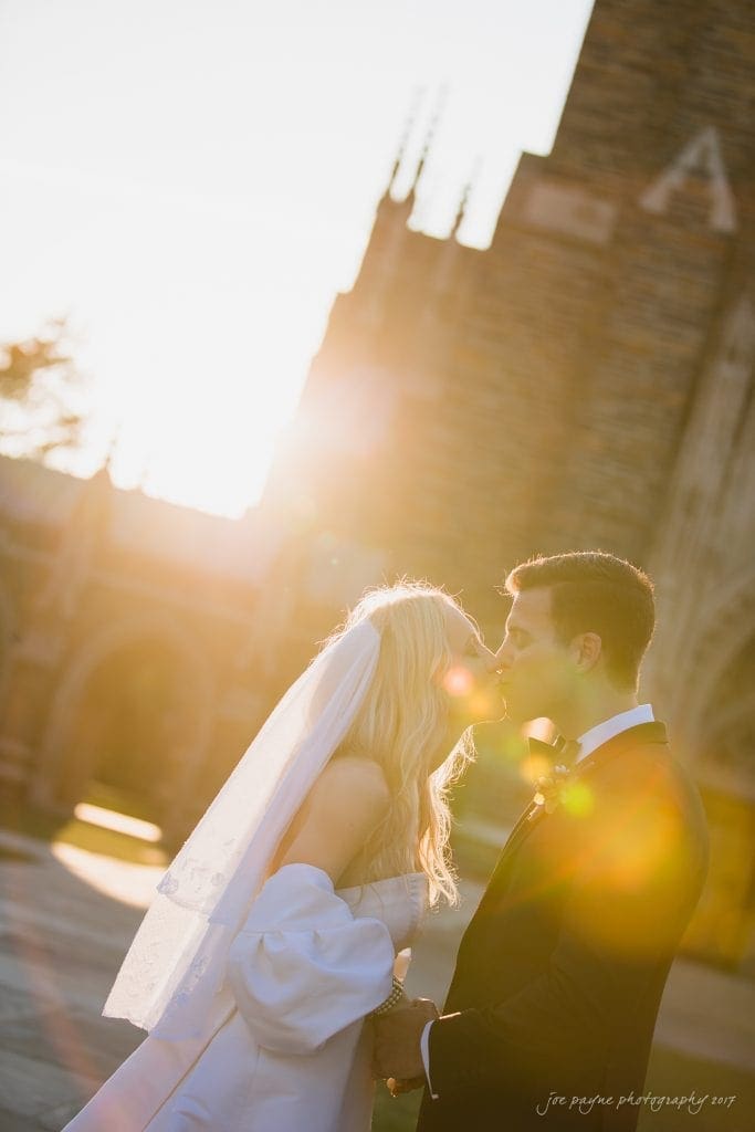 A romantic golden hour wedding portrait featuring a sun-flare kissed moment between the couple, with the warm glow illuminating their embrace against the intricate stone details of Duke Chapel in the background.