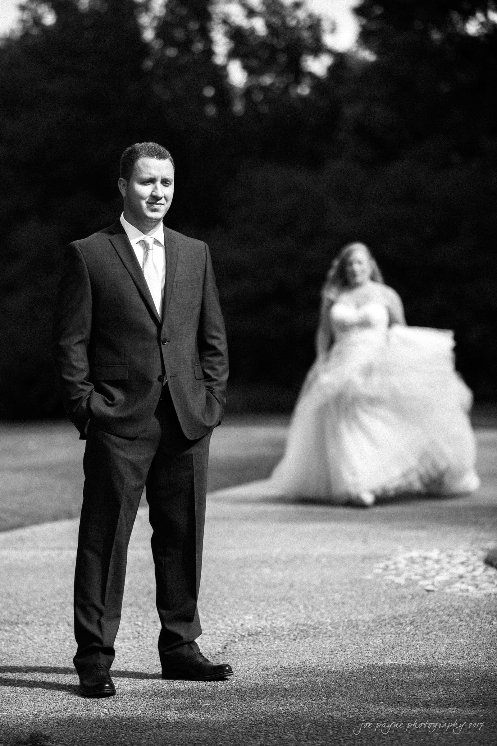 A groom in a suit stands with hands in pockets on a pathway, smiling slightly. In the background, a bride in a flowing white dress approaches, also smiling, with trees providing a leafy backdrop. The image is in black and white.