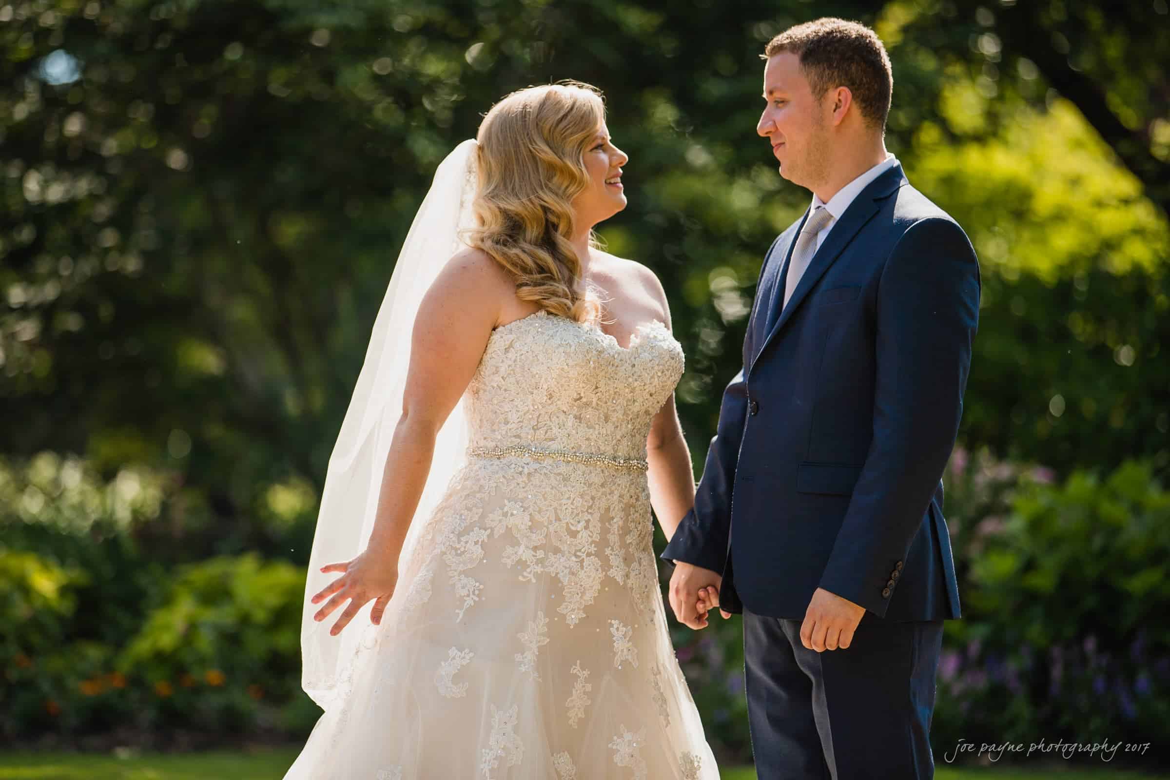A bride in a white lace gown and veil holds hands with a groom in a dark suit. They are standing outdoors in a lush garden setting, looking at each other fondly, with sunlight streaming through the trees.