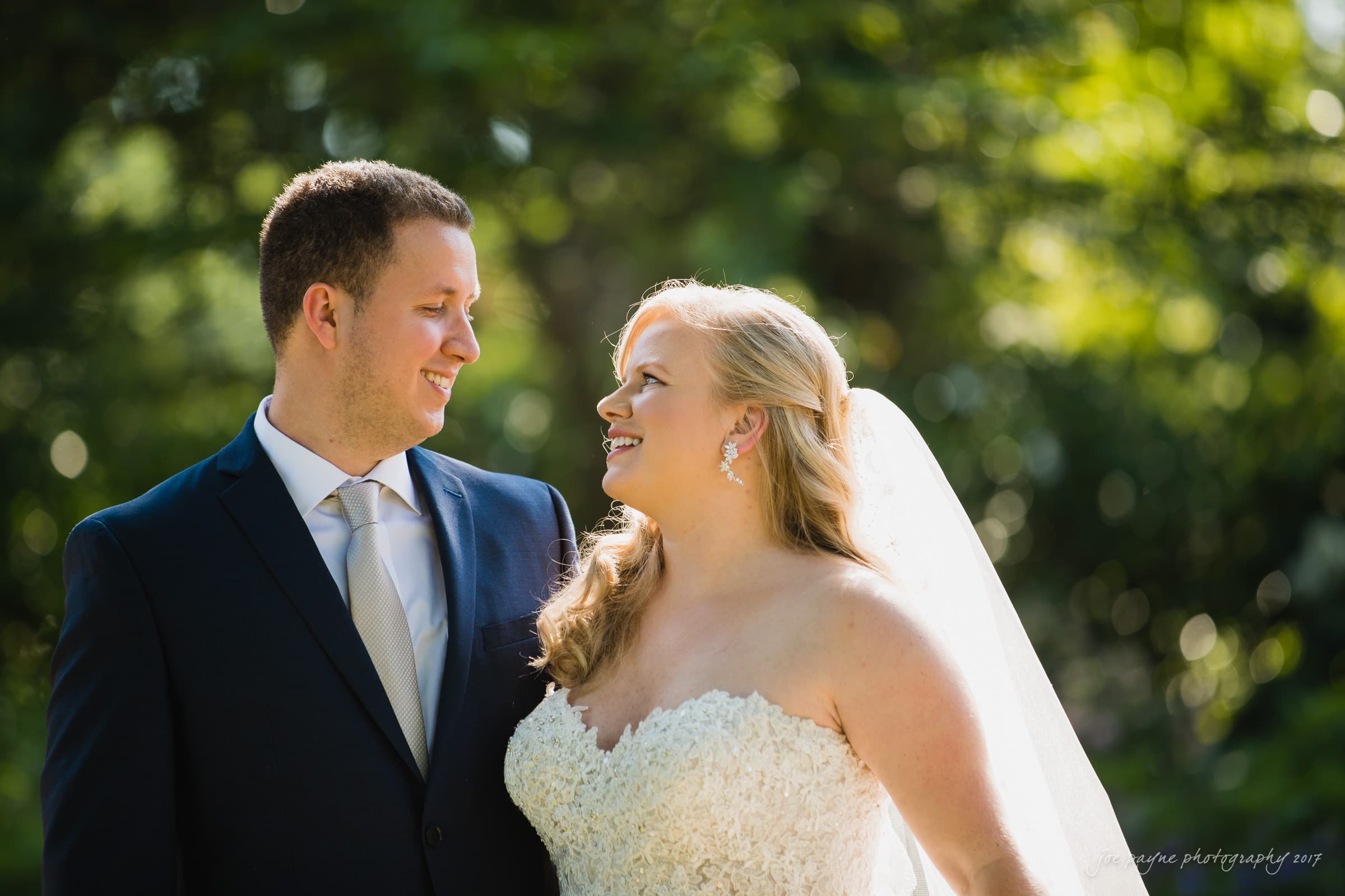 A bride and groom stand together outdoors. The bride is wearing a white lace dress and veil, while the groom is in a dark suit with a light tie. They are smiling at each other against a backdrop of greenery.
