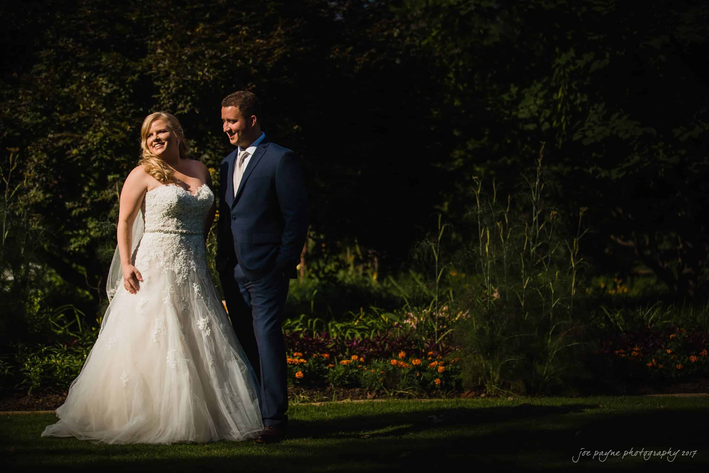 A bride and groom stand in a sunlit garden, with the bride wearing a white gown and the groom in a navy suit. The backdrop features lush greenery and colorful flowers.