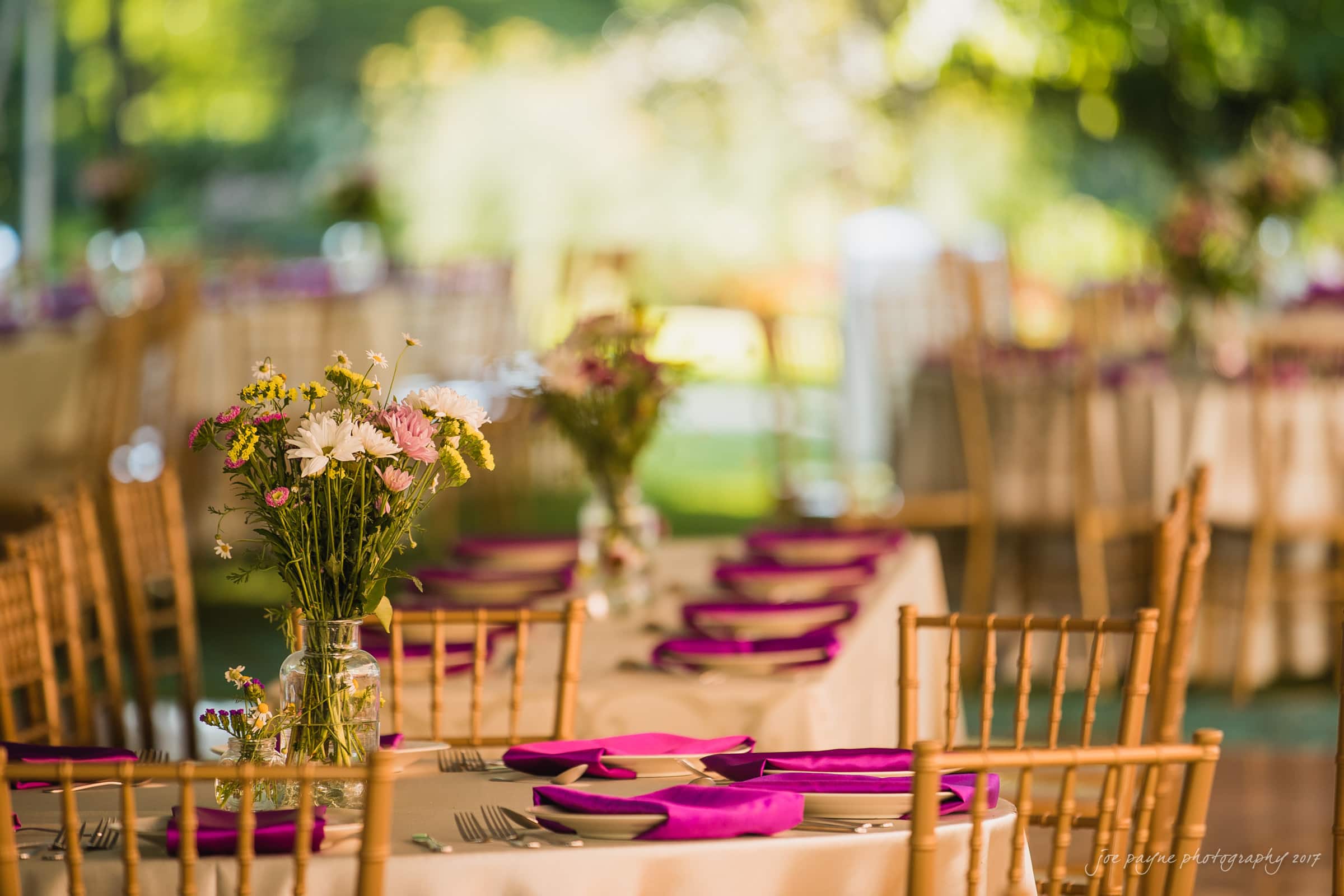 A beautifully set outdoor reception table adorned with pink napkins and floral centerpieces in glass vases. Wooden chairs surround the table, and the background features greenery and additional tables, creating a warm, inviting atmosphere.