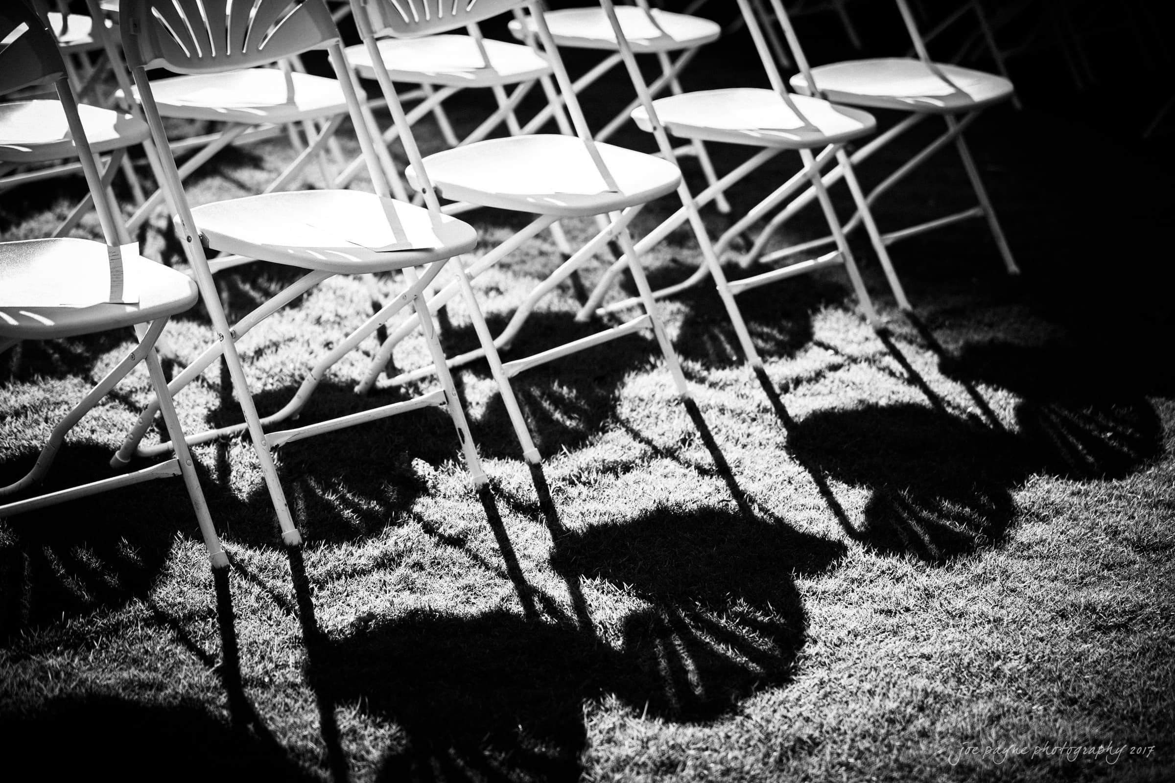 Black and white image of folding chairs arranged in rows on a lawn. The sunlight casts intricate shadows of the chair backs onto the grass, creating a pattern.