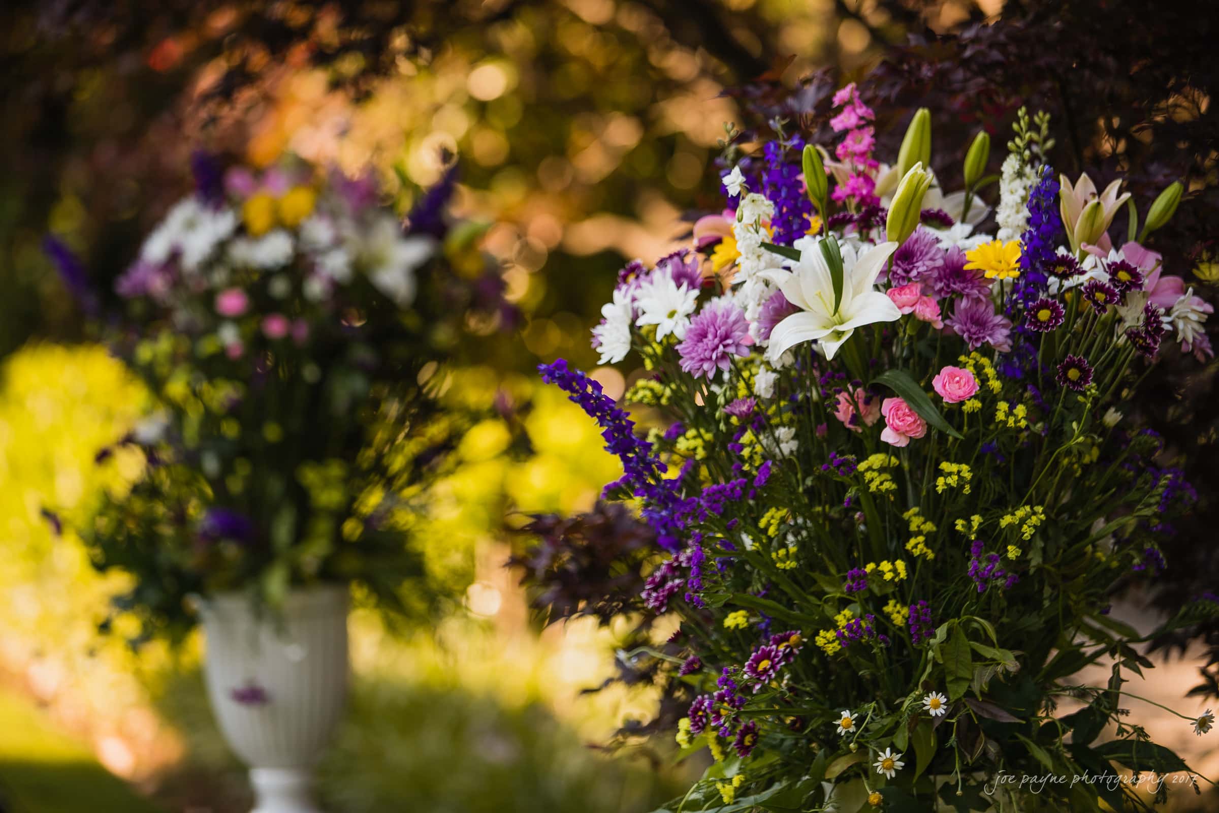 A vibrant floral arrangement featuring lilies, daisies, and various colorful wildflowers in a white vase, set against a blurred outdoor garden backdrop with warm lighting.
