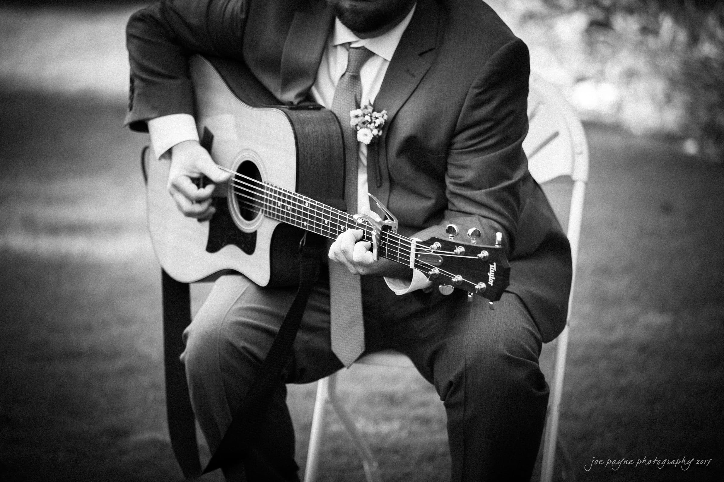 A man in a suit plays an acoustic guitar while sitting on a white folding chair outdoors. The image is black and white, capturing a moment of musical performance in a natural setting.
