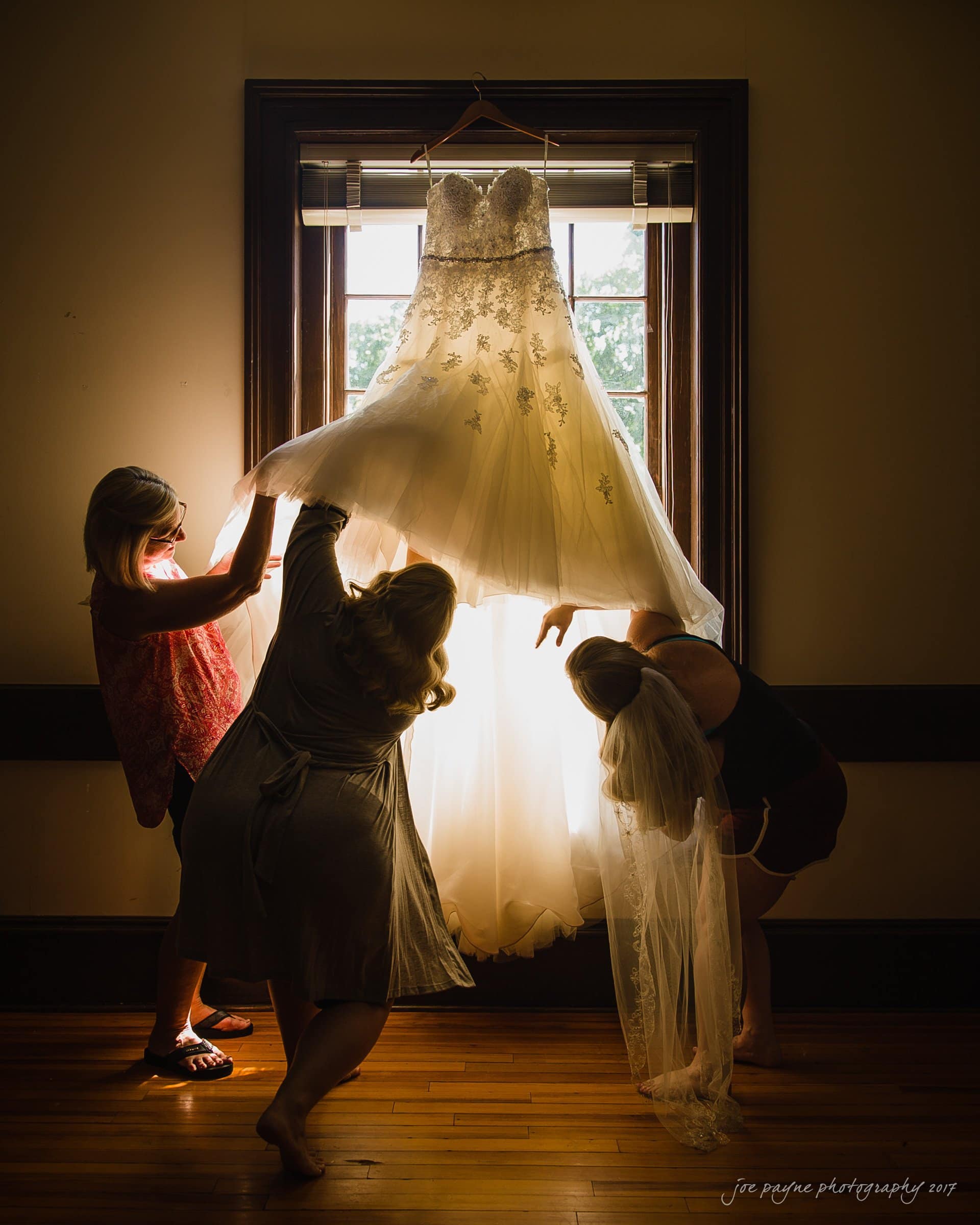 Three women hold up a wedding dress in front of a window, with sunlight streaming through the fabric, creating a glowing effect. The room features wooden floors and a framed window in the background.