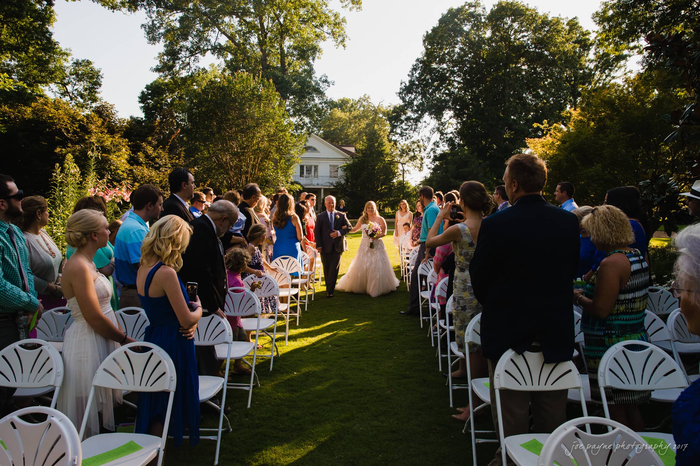 A bride in a white gown walks down an outdoor aisle lined with guests seated on white chairs. The ceremony takes place on a sunny day, with trees and a house in the background. The guests wear colorful outfits.