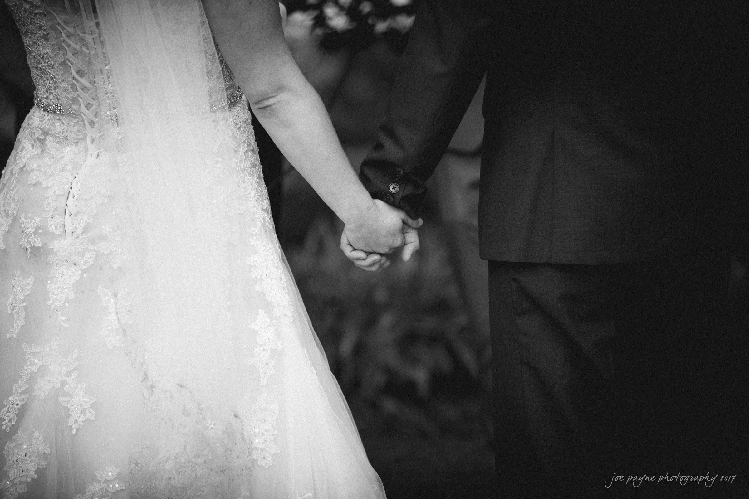 A black and white photo showing a bride and groom from behind, holding hands. The bride wears a lace wedding dress with a veil, while the groom is in a dark suit. The romantic scene is set outdoors.