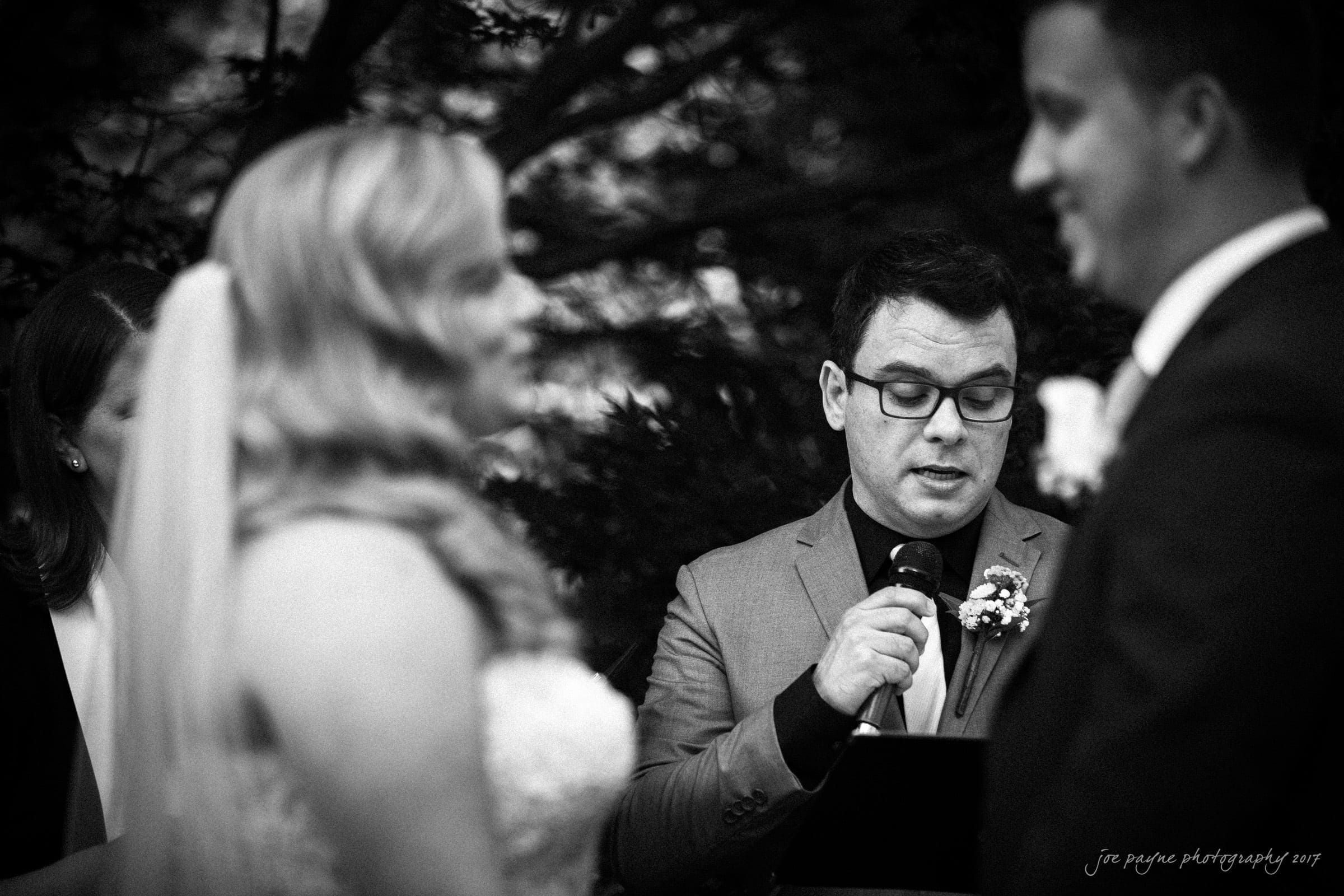 A black and white photo of a wedding ceremony. A man in glasses, holding a microphone, is speaking between a bride in a veil and a groom. They are outdoors with foliage in the background.