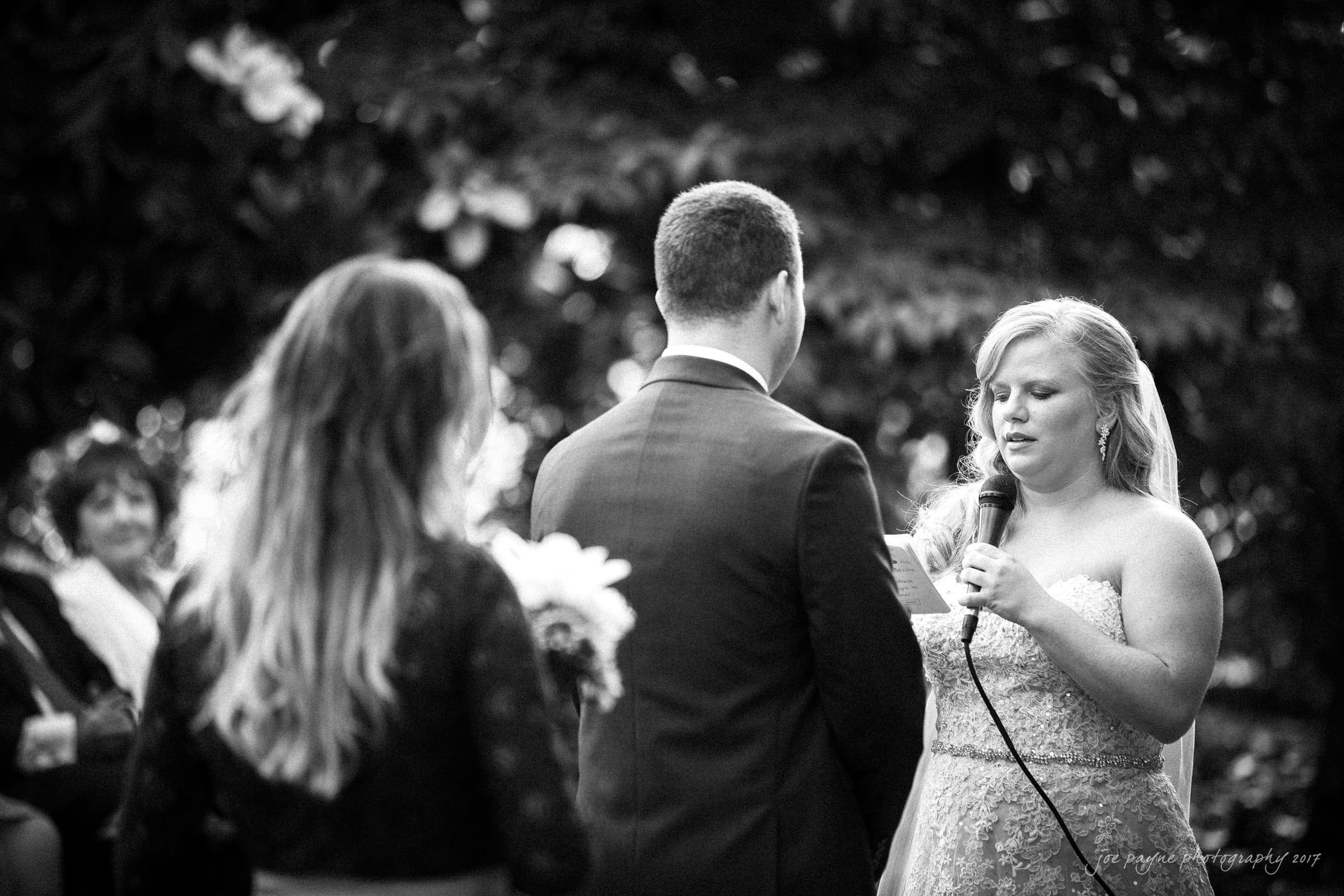 A bride and groom stand face to face during an outdoor wedding ceremony. The bride is speaking into a microphone, and another woman stands nearby holding a small bouquet. Guests are seated in the background.