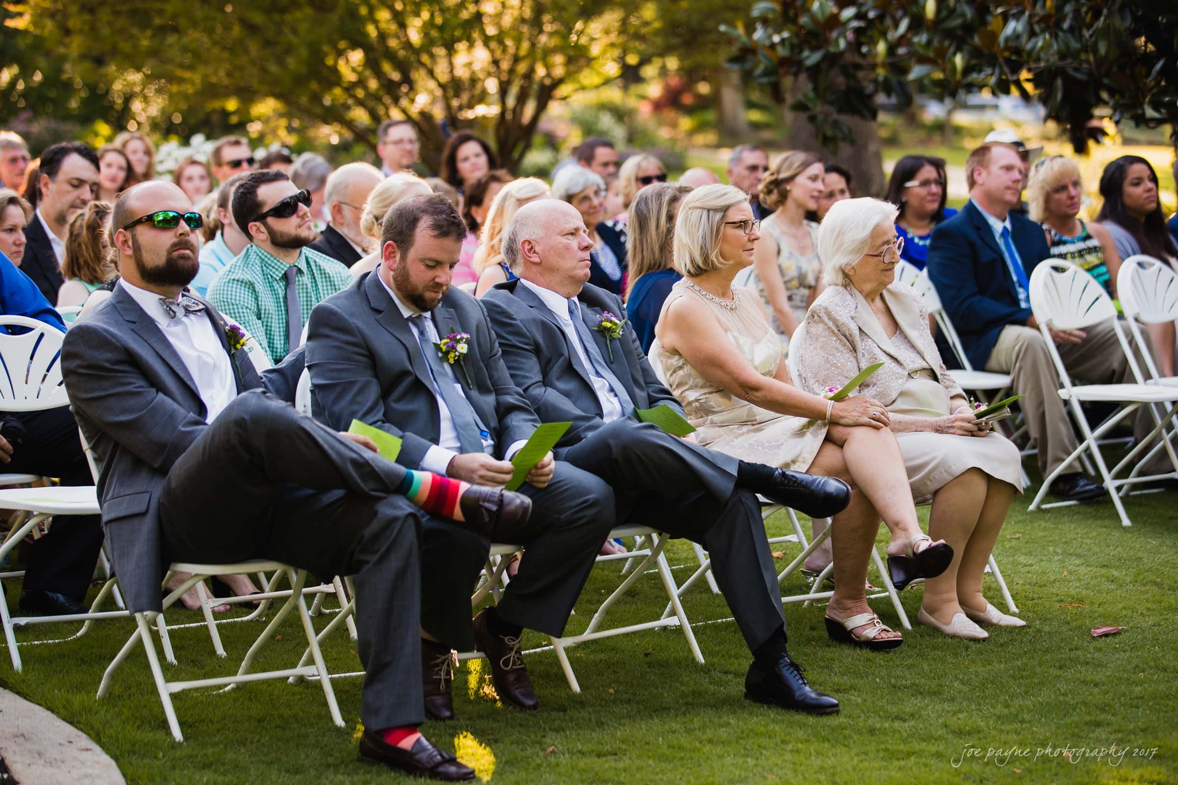 A group of people seated outdoors at an event, with men and women dressed formally. Some hold papers, and one man wears vibrant striped socks. The setting is lush with trees and greenery in the background.