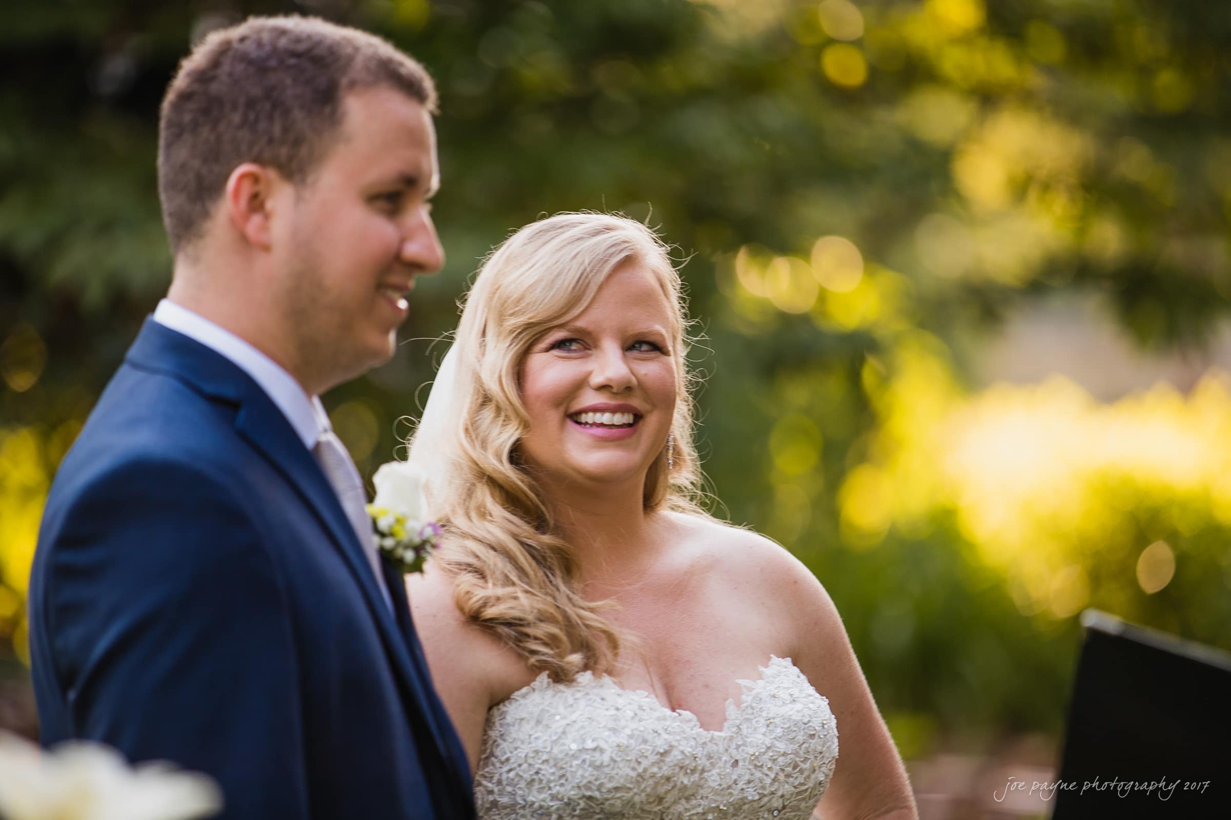 A bride and groom are smiling outdoors in a lush green setting. The bride is wearing a strapless white gown and veil, while the groom is in a dark suit with a white shirt. They appear to be attending a ceremony or celebration.