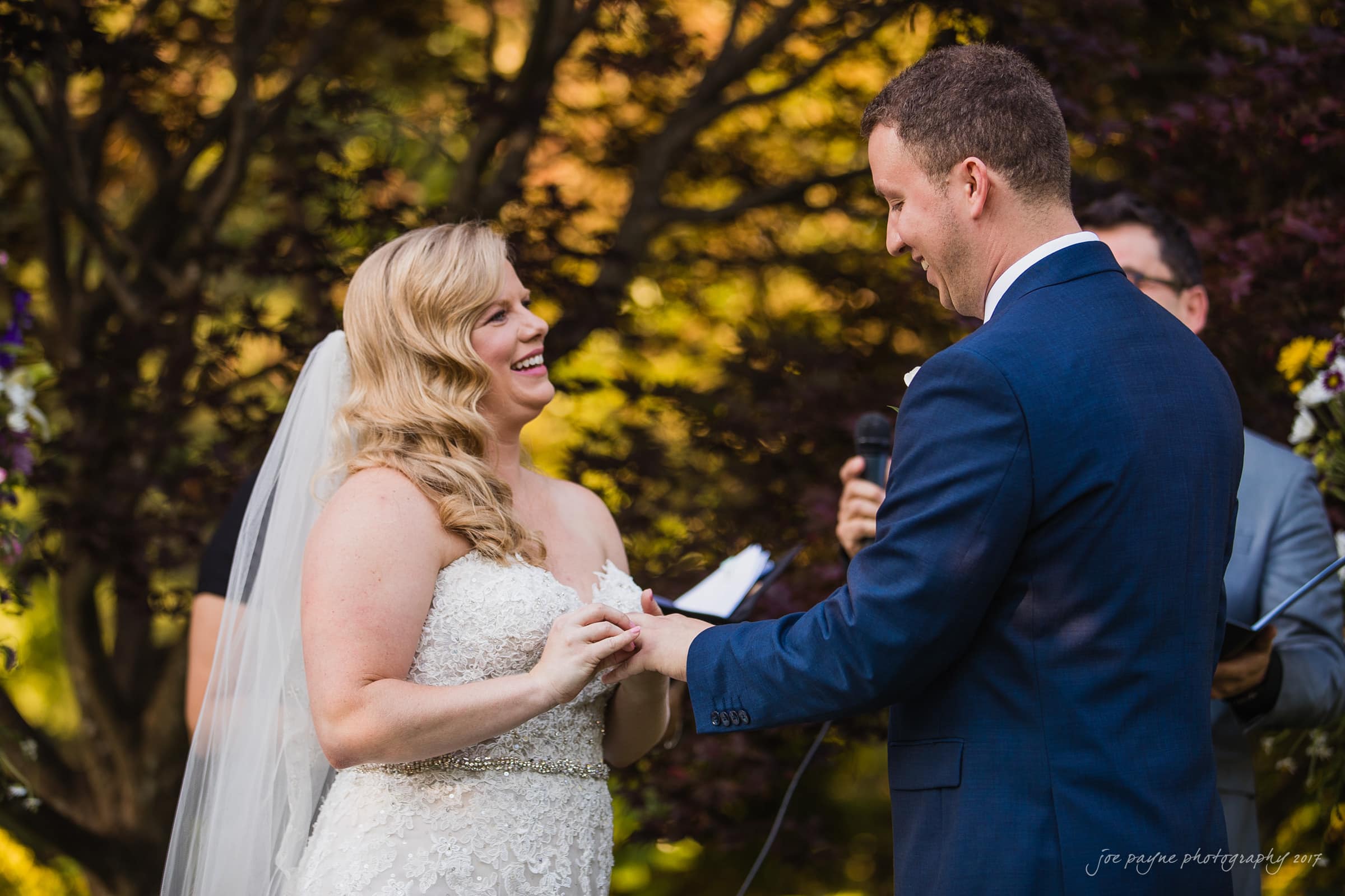 A bride and groom stand facing each other outdoors during their wedding ceremony. The bride is placing a ring on the grooms finger. Both are smiling warmly. The background is filled with green and orange foliage.