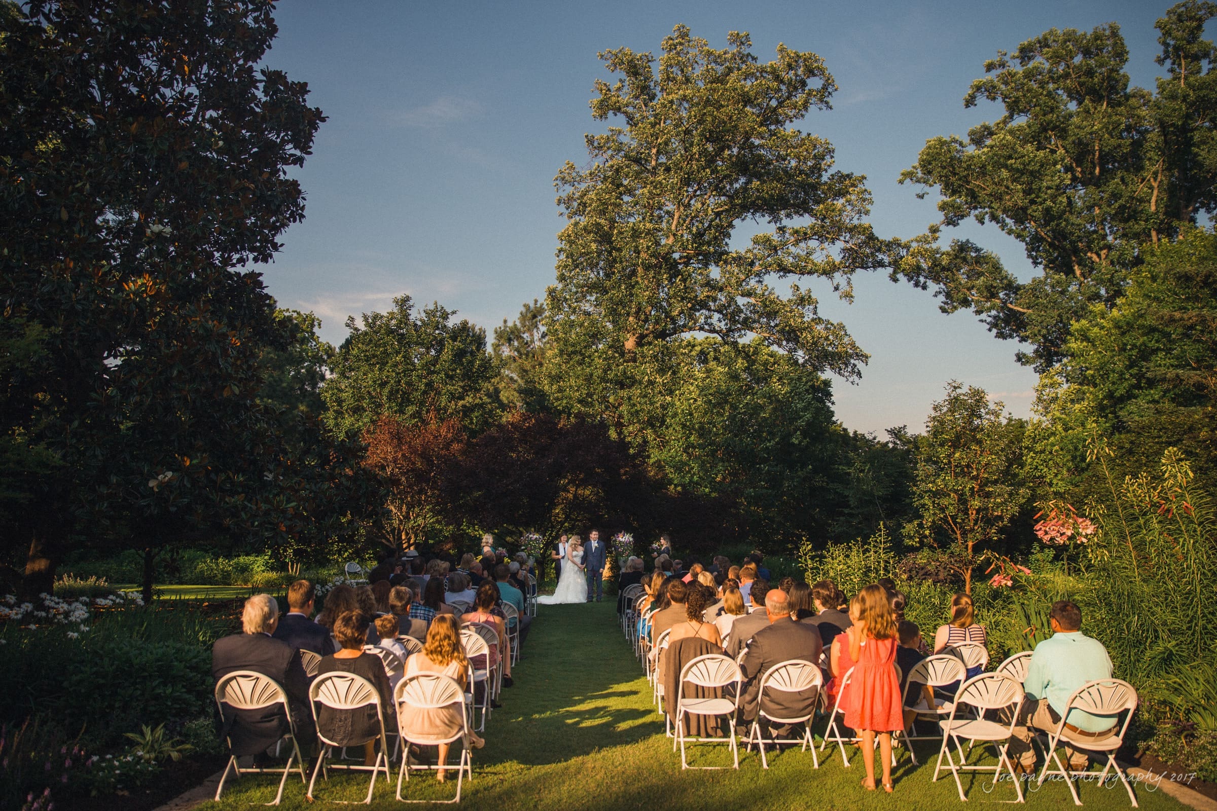An outdoor wedding ceremony in a garden setting on a sunny day. Guests are seated on white chairs, facing a couple standing at the altar. Large trees and lush greenery surround the scene. A child in a bright orange dress stands among the guests.