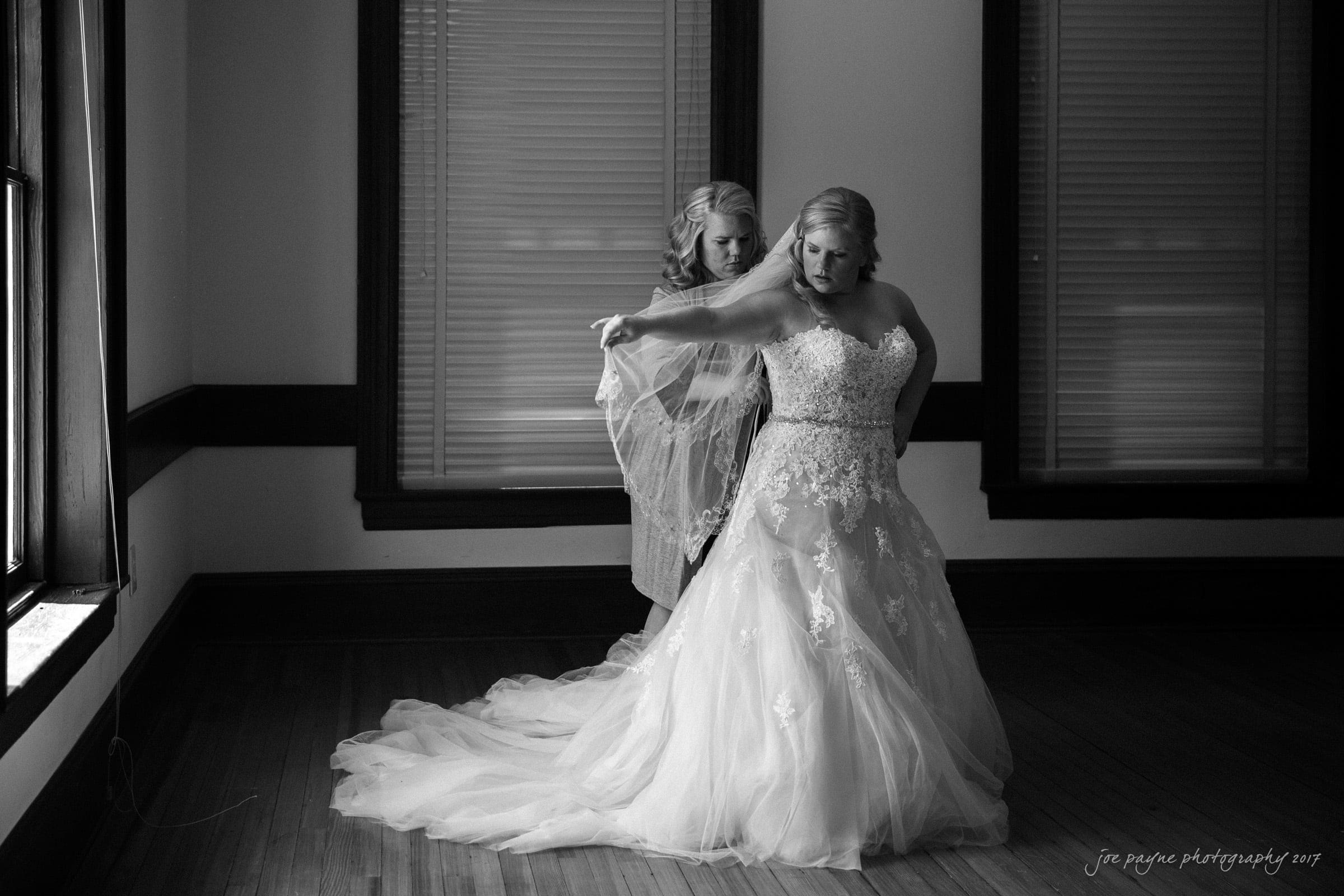 A bride stands in a gown with intricate lace detailing, while another woman assists her with a veil. They are in a room with large windows and wooden floors. The scene is in black and white, capturing an intimate and elegant moment.