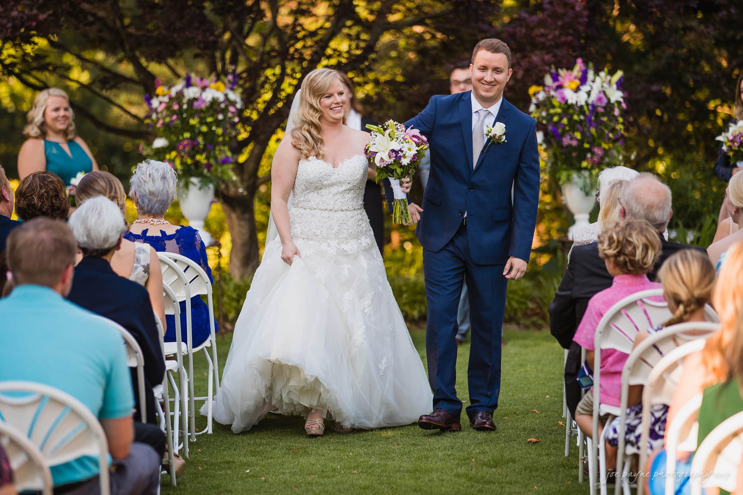 A bride and groom smile while walking down an outdoor aisle, surrounded by guests seated on white chairs. The groom wears a blue suit, and the bride wears a white gown, holding a bouquet. Lush greenery and floral arrangements are visible in the background.