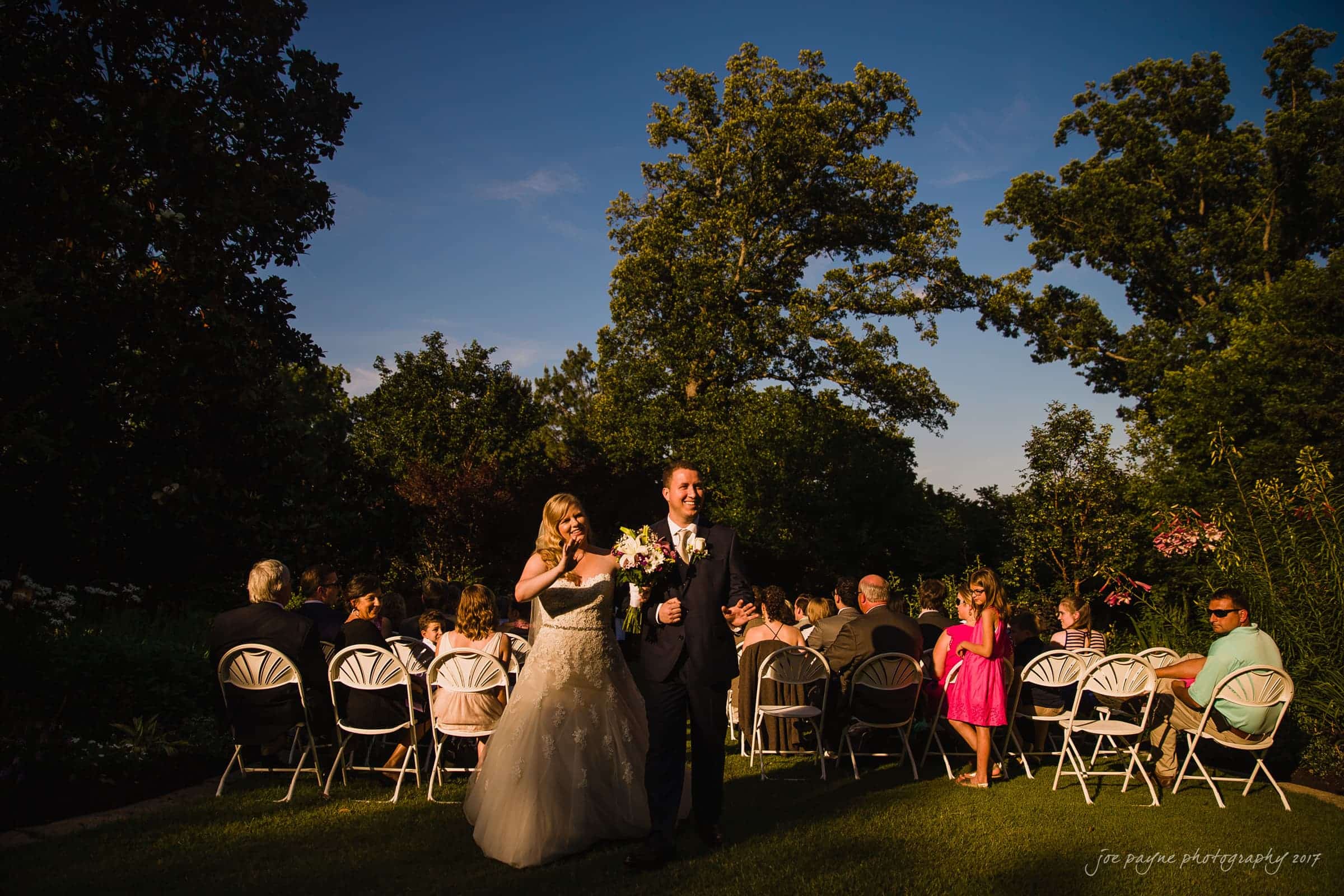 A newlywed couple joyfully walks down the aisle outdoors, surrounded by seated guests in white chairs. The bride holds a bouquet, and the groom wears a suit. Trees and clear blue sky form the backdrop.