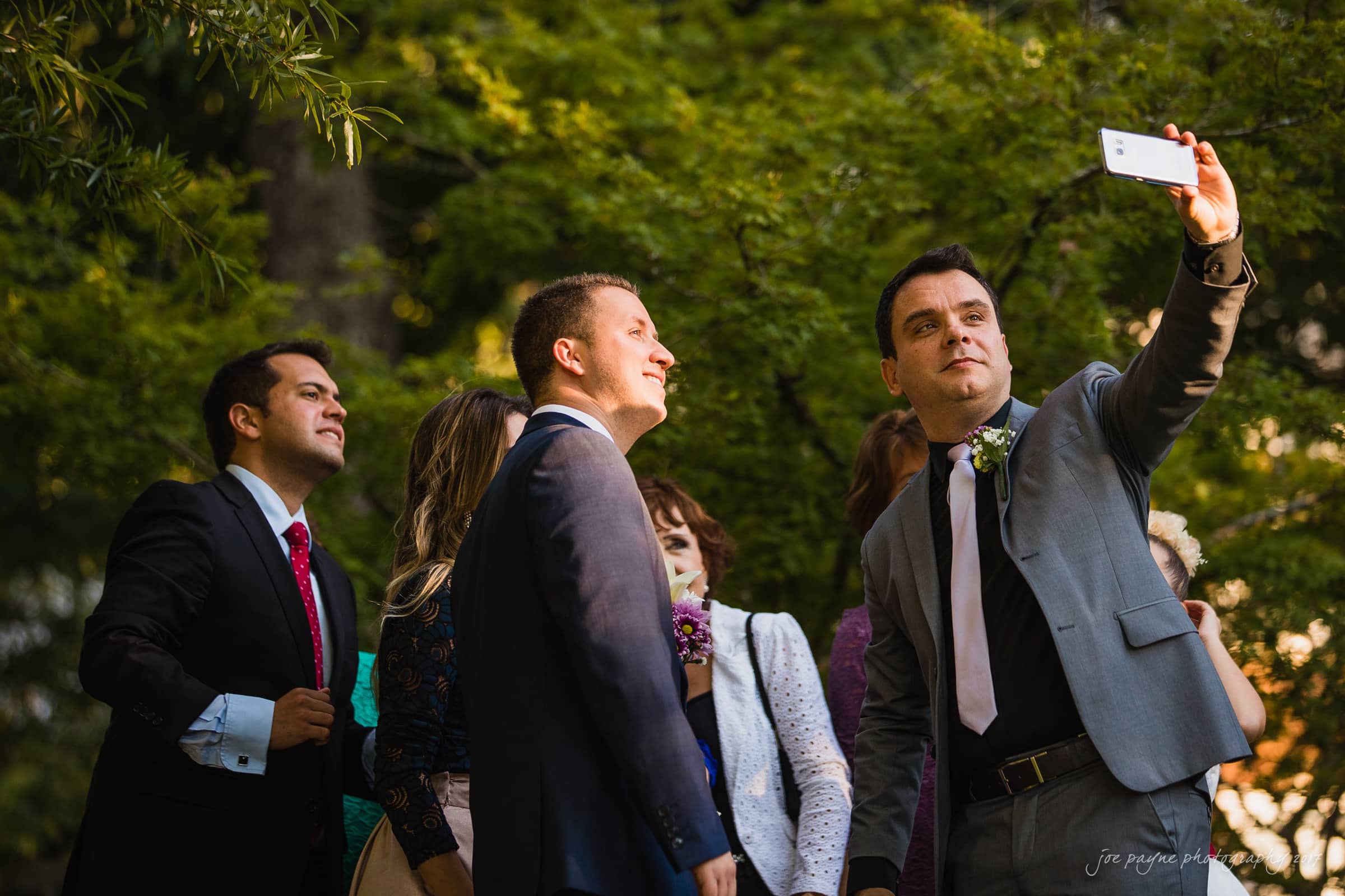 A group of people in formal attire are outdoors, posing for a selfie. The person in the front right is holding a smartphone to capture the moment. Green foliage is visible in the background, suggesting a garden or park setting.